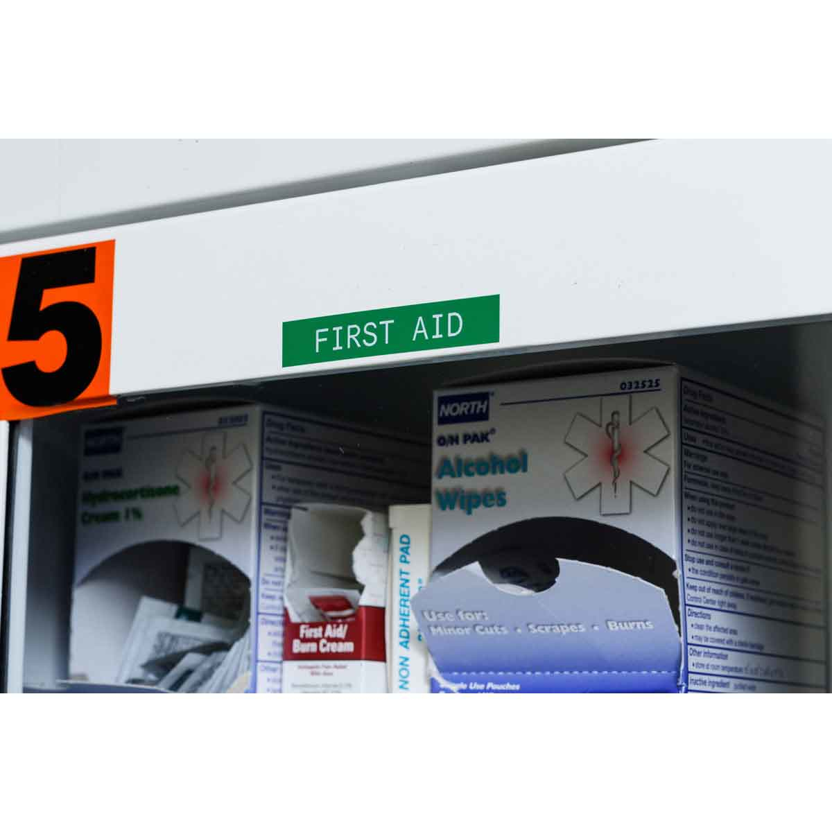 Interno di un armadietto di pronto soccorso. Sono visibili forniture mediche e un cartello verde con la scritta 'FIRST AID'.