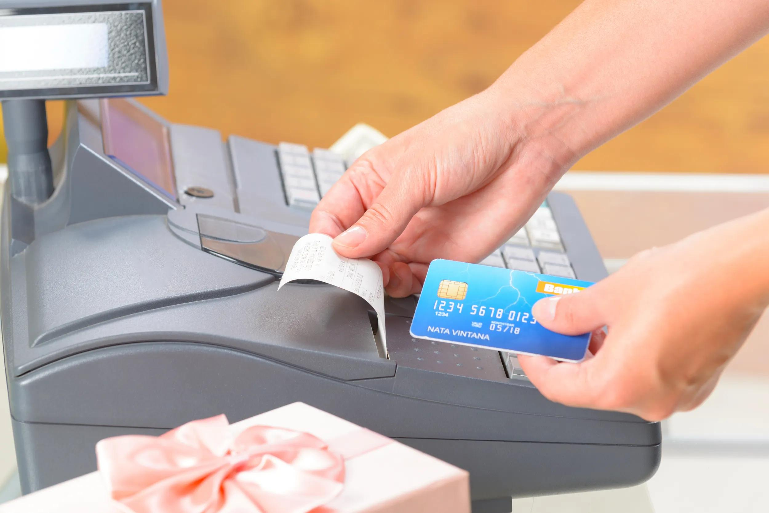 Una mano che tiene una carta di credito e una ricevuta vicino a un registratore di cassa, con un pacco regalo.