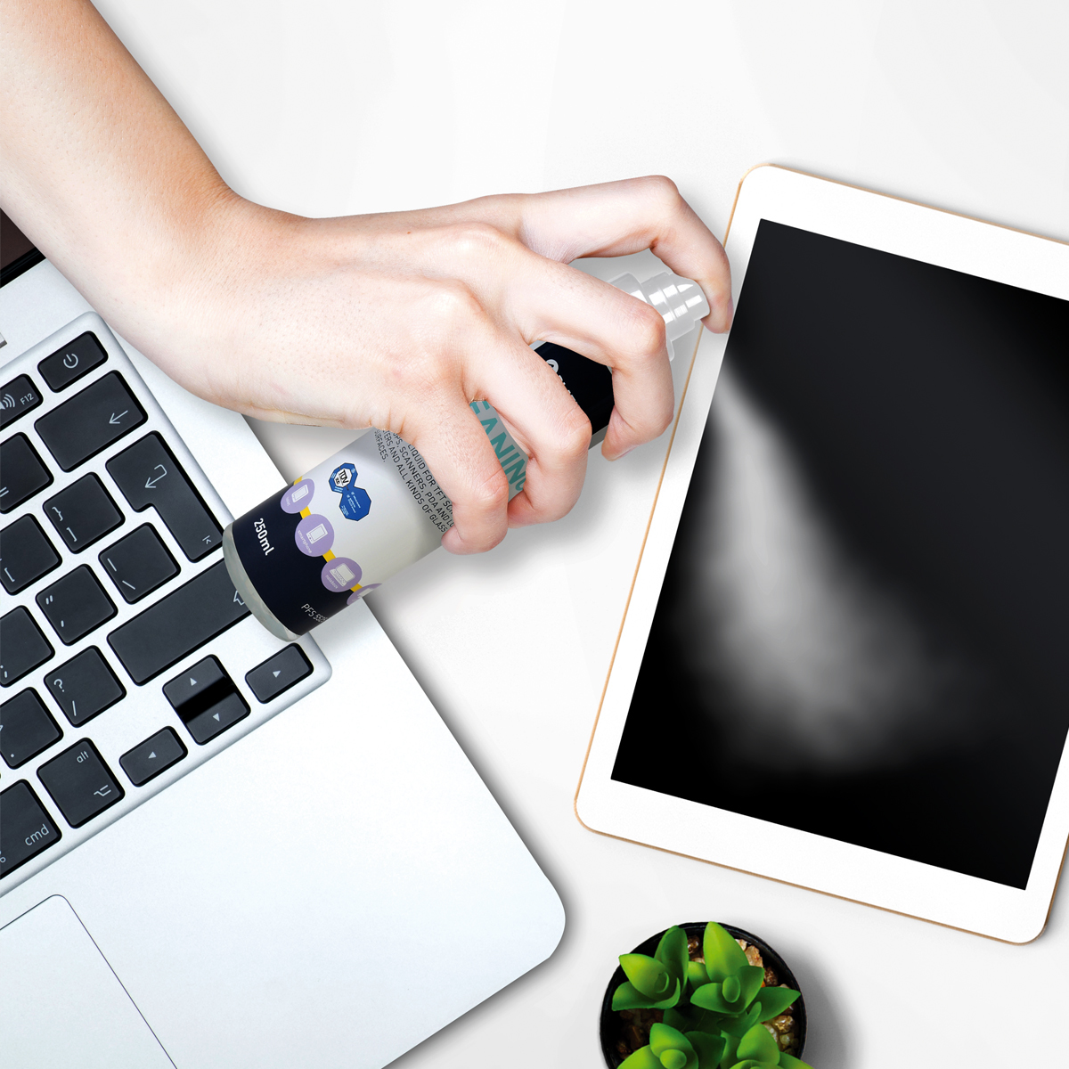 Una mano spruzza una soluzione detergente su un laptop e un tablet, su una superficie bianca.