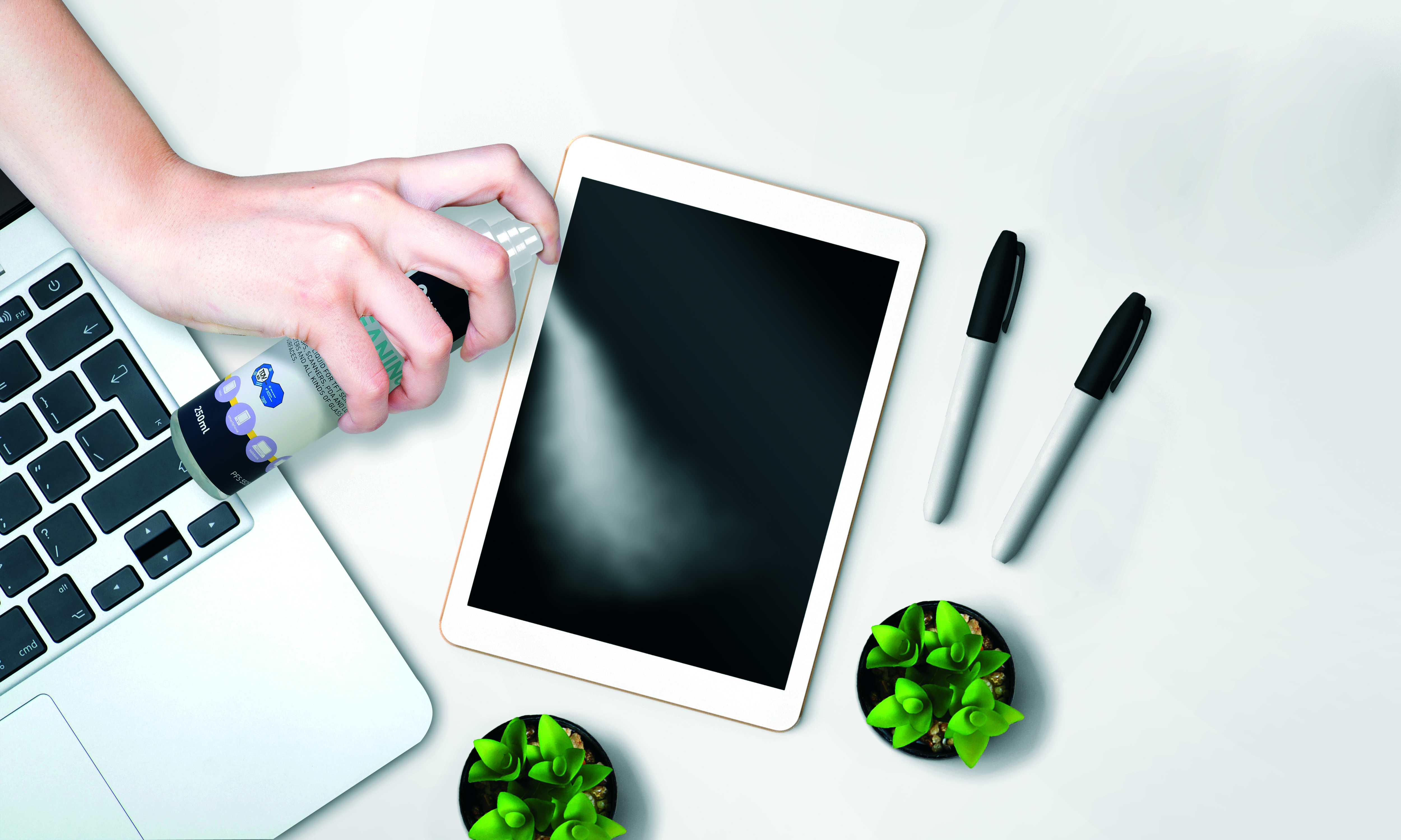 Una mano che spruzza una soluzione detergente su un tablet bianco. Un laptop e pennarelli sono anche sulla superficie bianca.