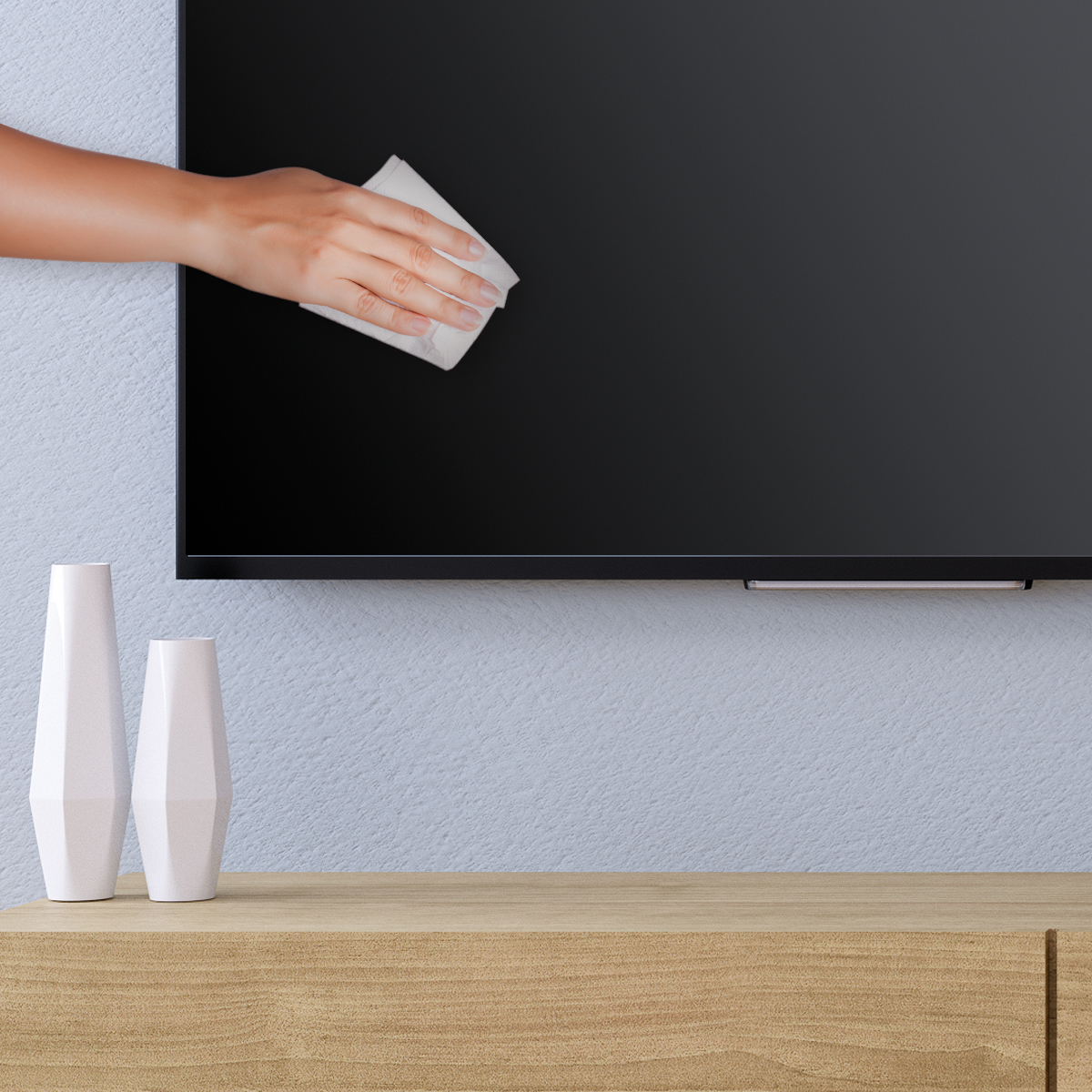 Una mano pulisce uno schermo TV nero con un panno bianco. Due vasi bianchi sono su una superficie di legno.