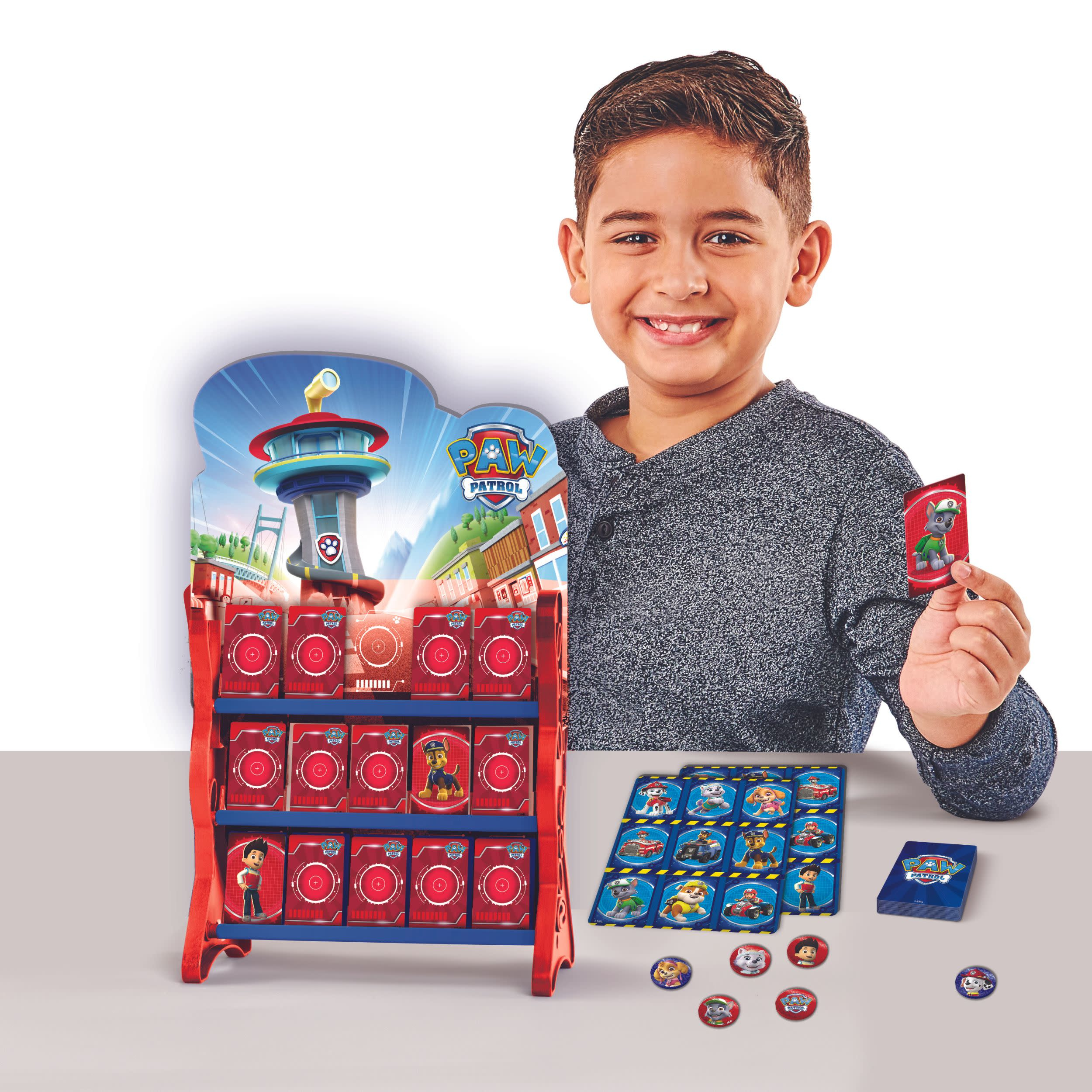 Ein Junge hält eine Karte von einem PAW Patrol-Spiel, mit einer Spielanzeige und anderen Karten auf einem Tisch.