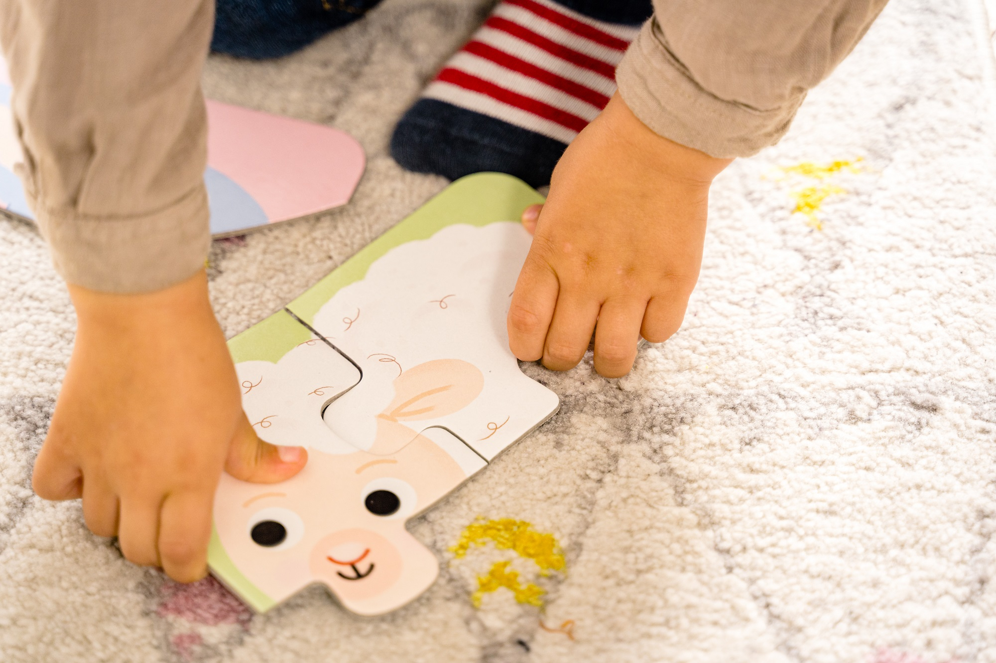 Ręce dziecka dopasowują do siebie puzzle z kreskówkową owcą na biało-szarym wzorzystym dywanie.