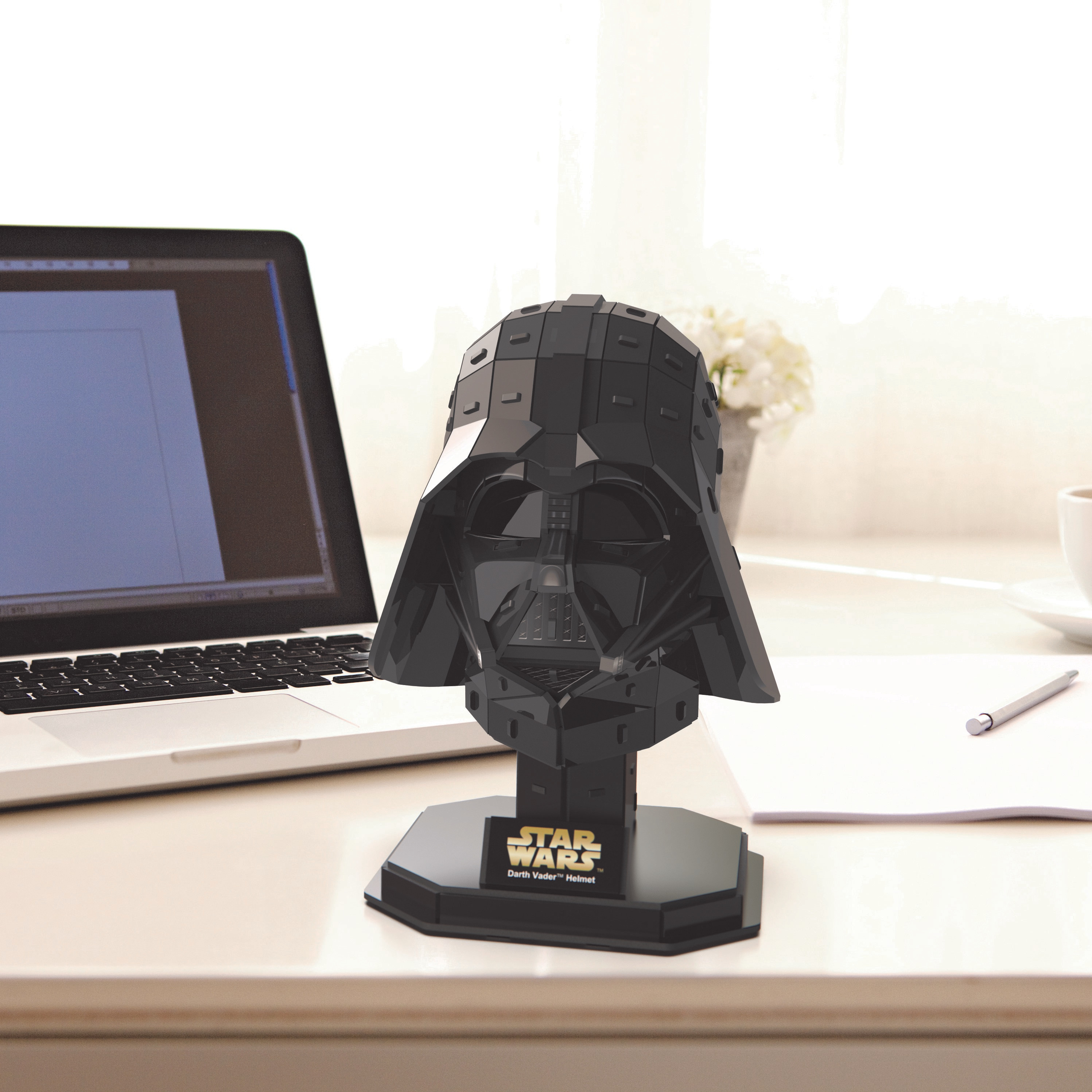 Darth Vader helmet puzzle on a desk. Black helmet, laptop, pen, and flowers in the background.