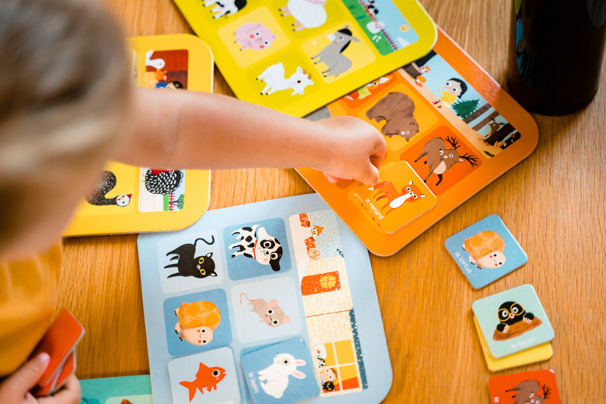 Dłoń dziecka sięgająca po kartę na stole z obrazkowymi kartami o tematyce zwierzęcej. Kilka kart jest rozrzuconych.