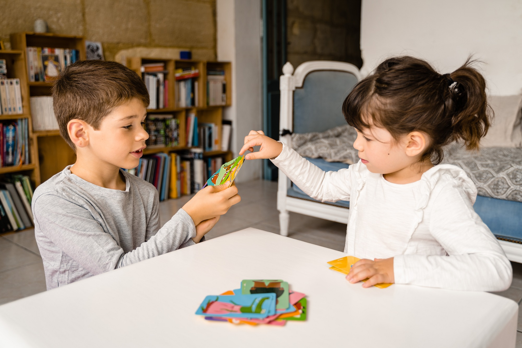 Dwoje dzieci gra w karty przy białym stole. Regały z książkami są w tle.