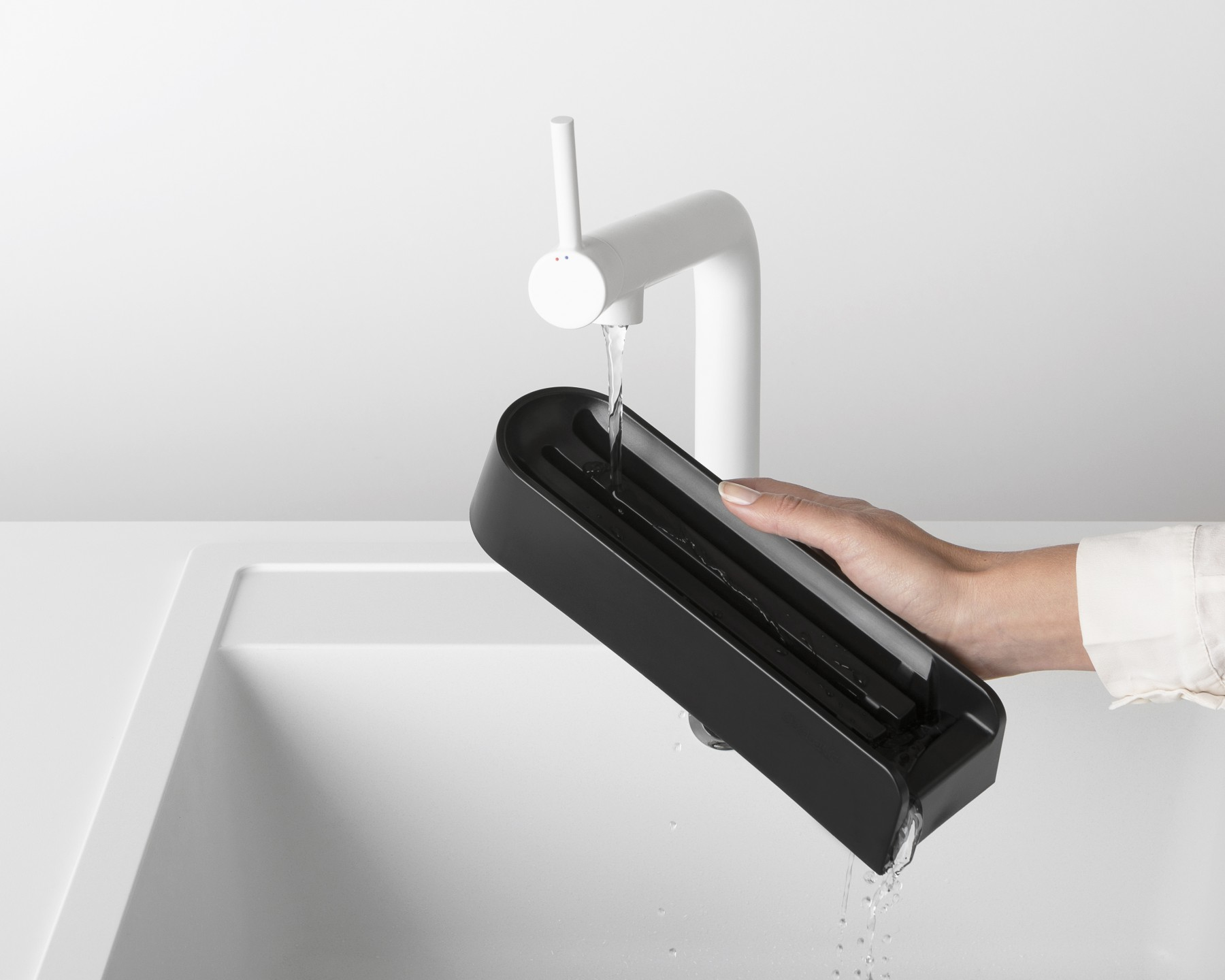 Eine Hand, die einen schwarzen Messerhalter unter einem weißen Wasserhahn hält, Wasser fließt ins Waschbecken.