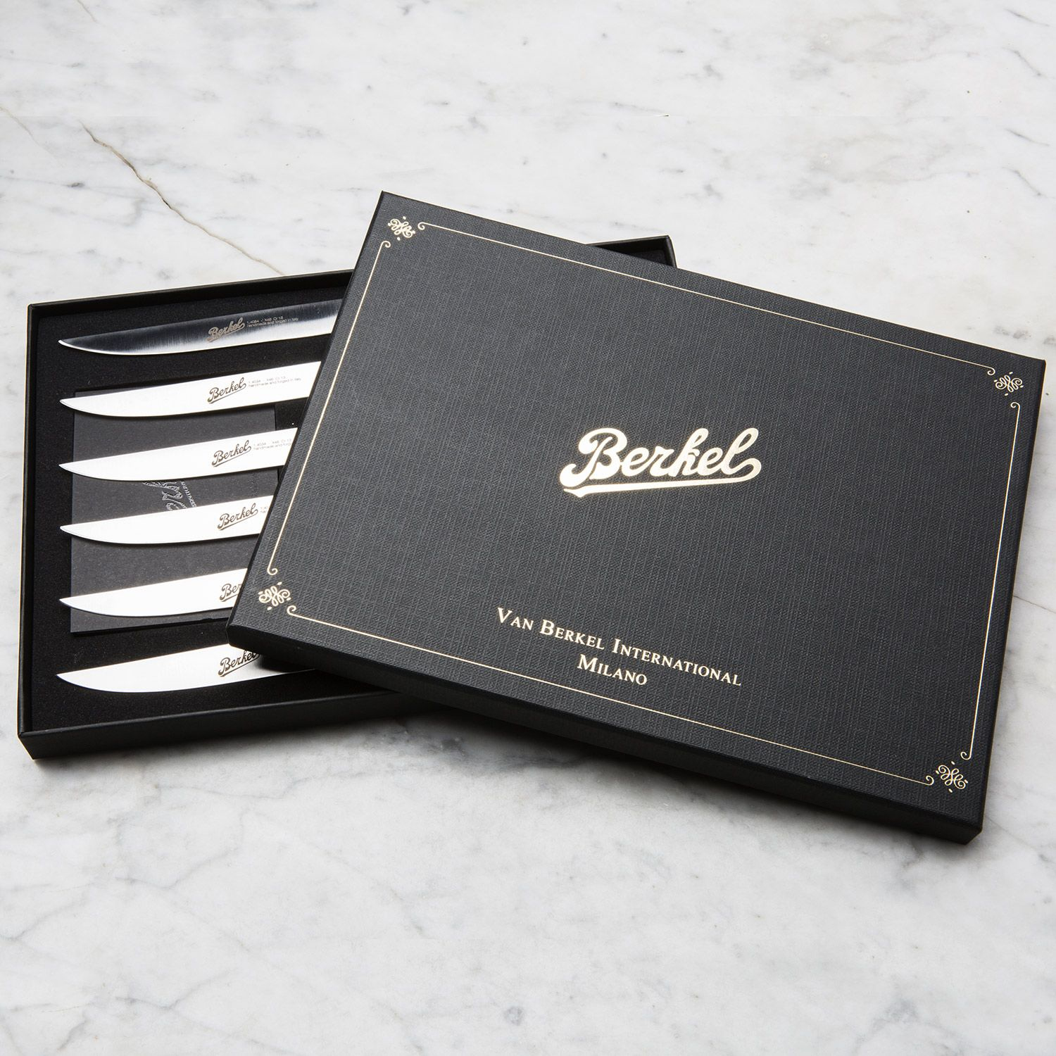 Czarne pudełko ze złotymi napisami i otwartą pokrywą, odsłaniające pięć noży ze srebrnymi ostrzami.