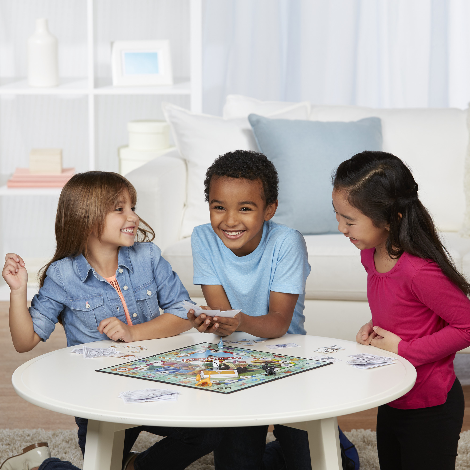 Drei Kinder spielen ein Brettspiel auf einem weißen Tisch und lächeln. Im Hintergrund ist eine Couch zu sehen.