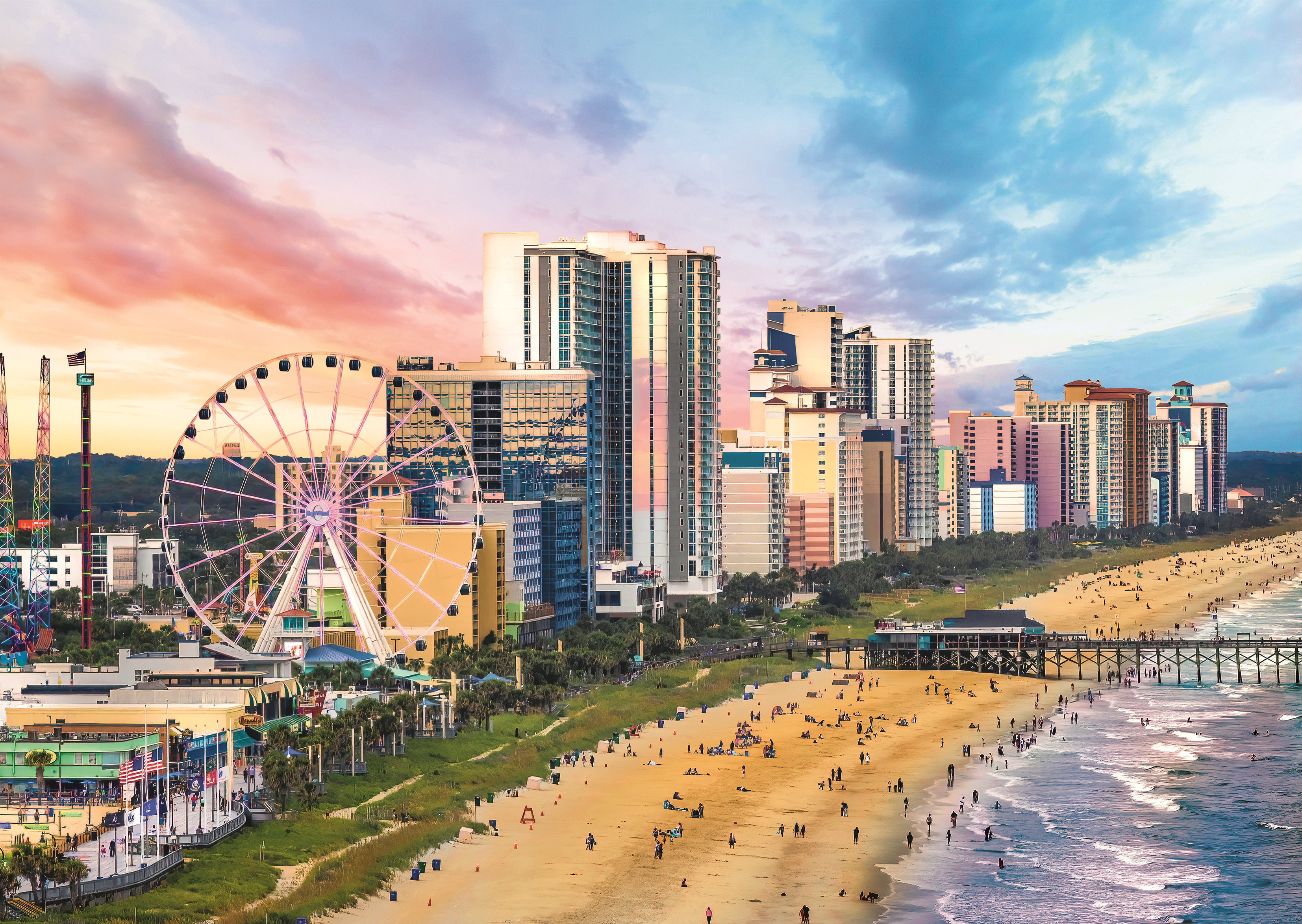 Una spiaggia con edifici alti, una ruota panoramica, persone e un molo al tramonto. Il cielo è rosa e blu.