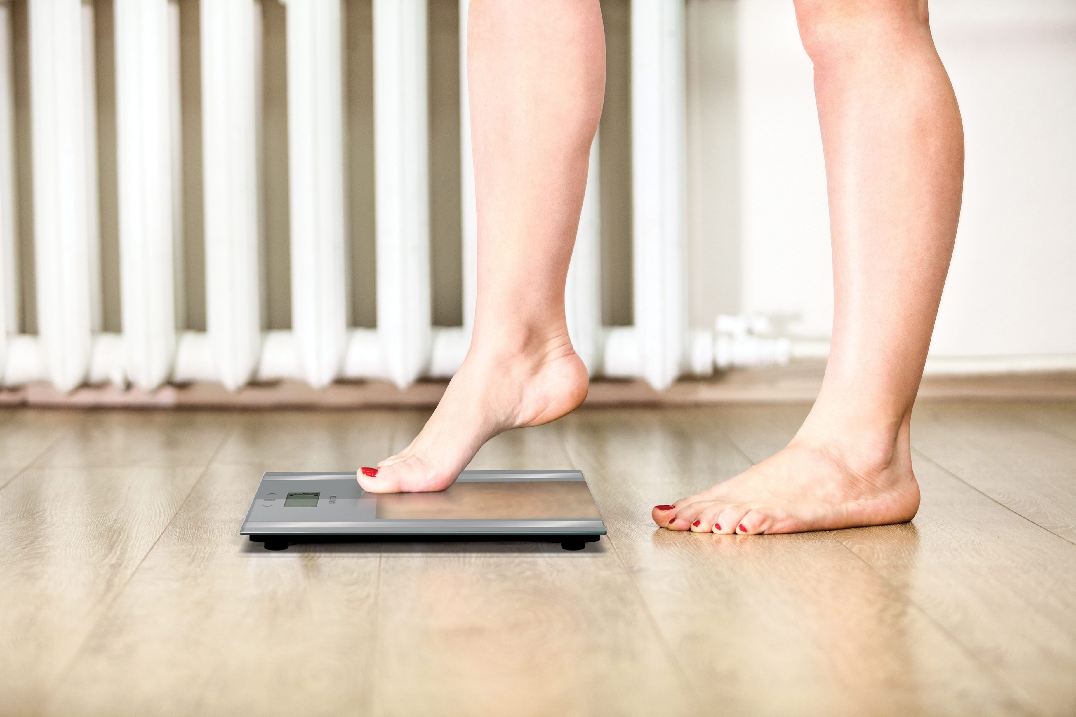 Stopa osoby na cyfrowej wadze łazienkowej. Waga wskazuje 0,0 kg, a podłoga jest drewniana.