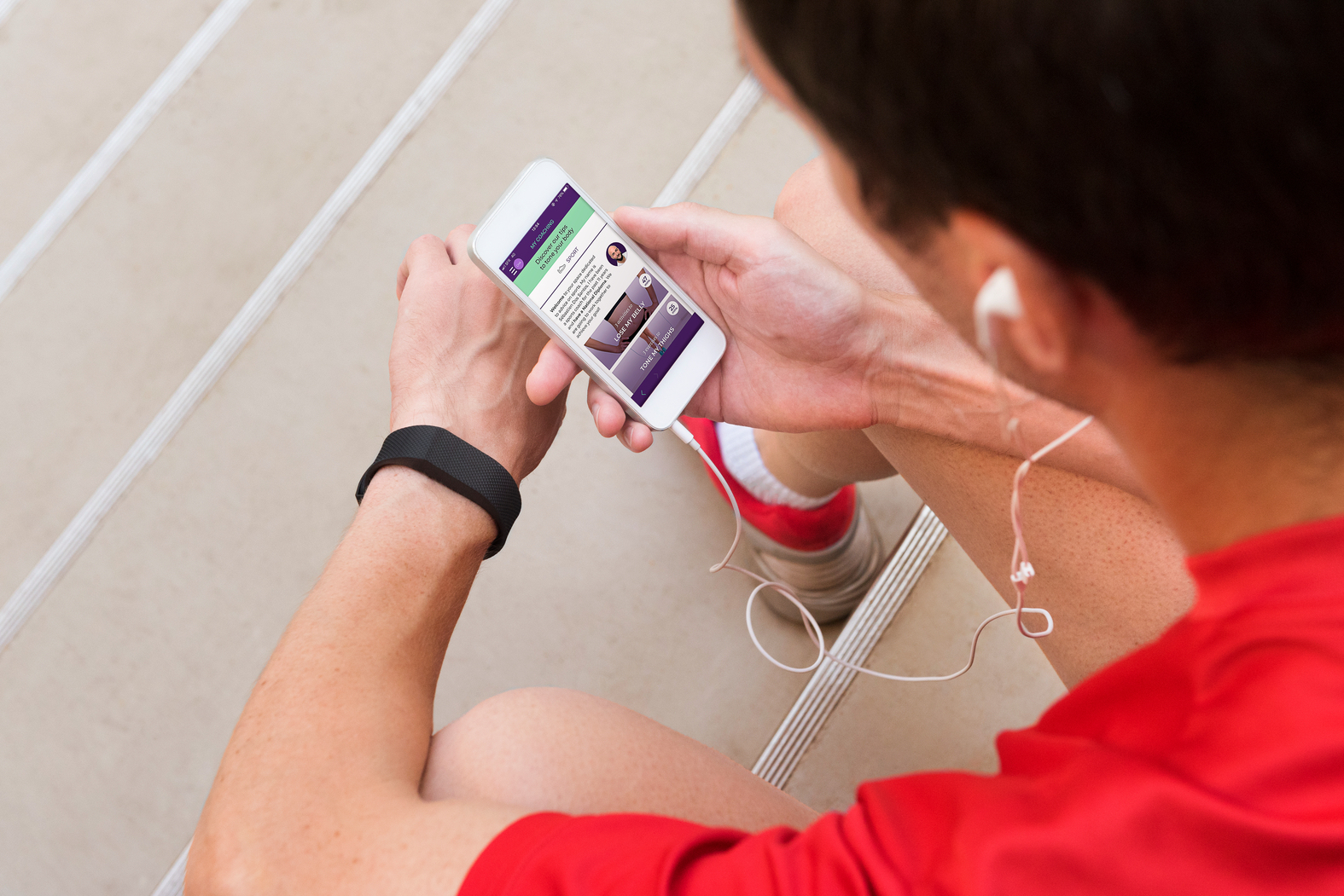 Mężczyzna siedzi na schodach, patrzy na ekran telefonu z aplikacją fitness. Słuchawki i zegarek widoczne.