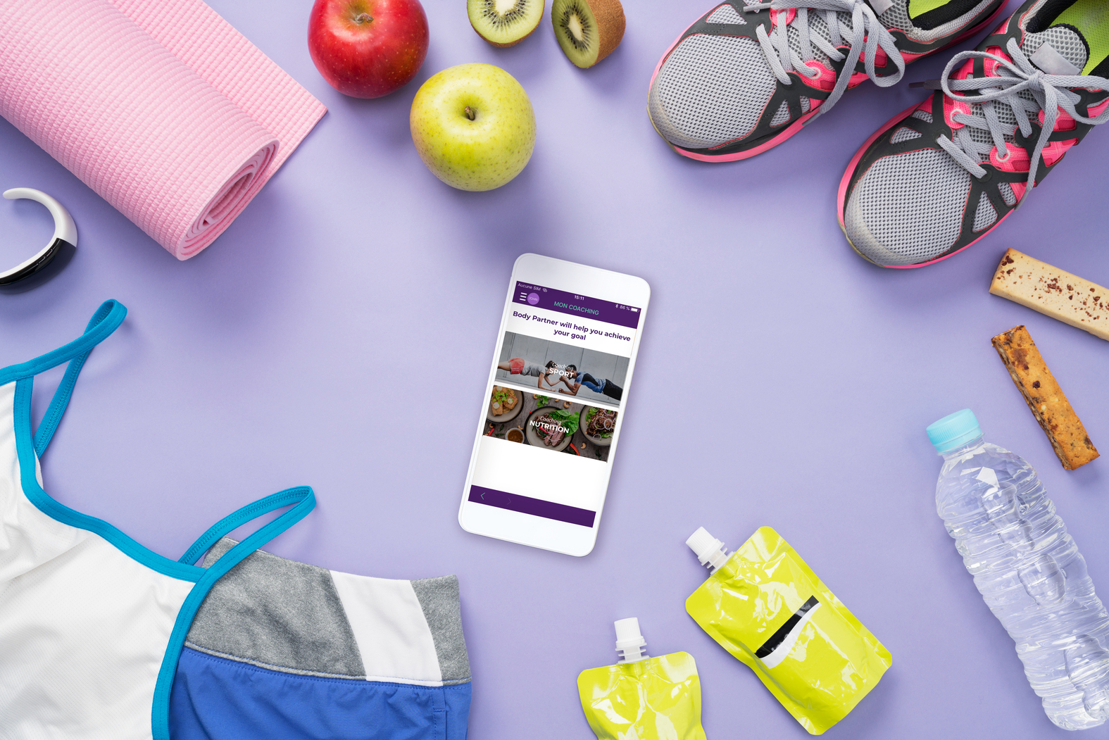 Przedmioty fitness: telefon z aplikacją, buty, mata, owoce, pakiety energetyczne na fioletowej powierzchni.
