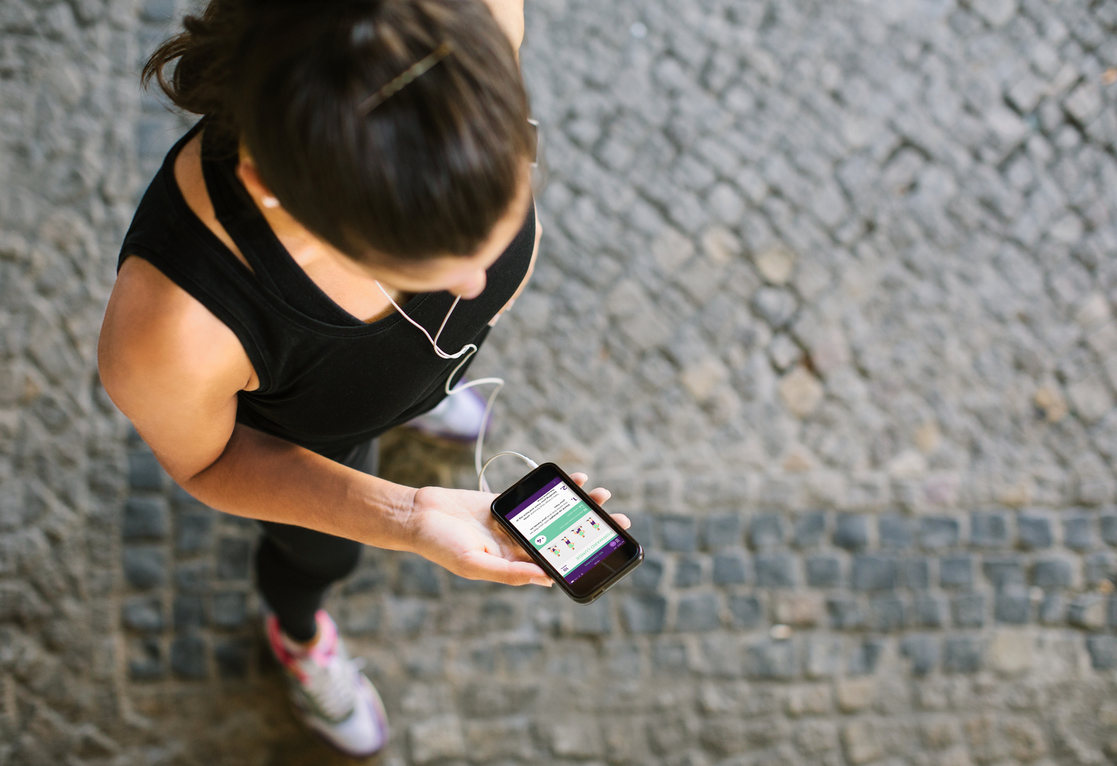 Kobieta biegnie, patrzy na ekran telefonu z aplikacją fitness. Widoczna brukowana ulica.