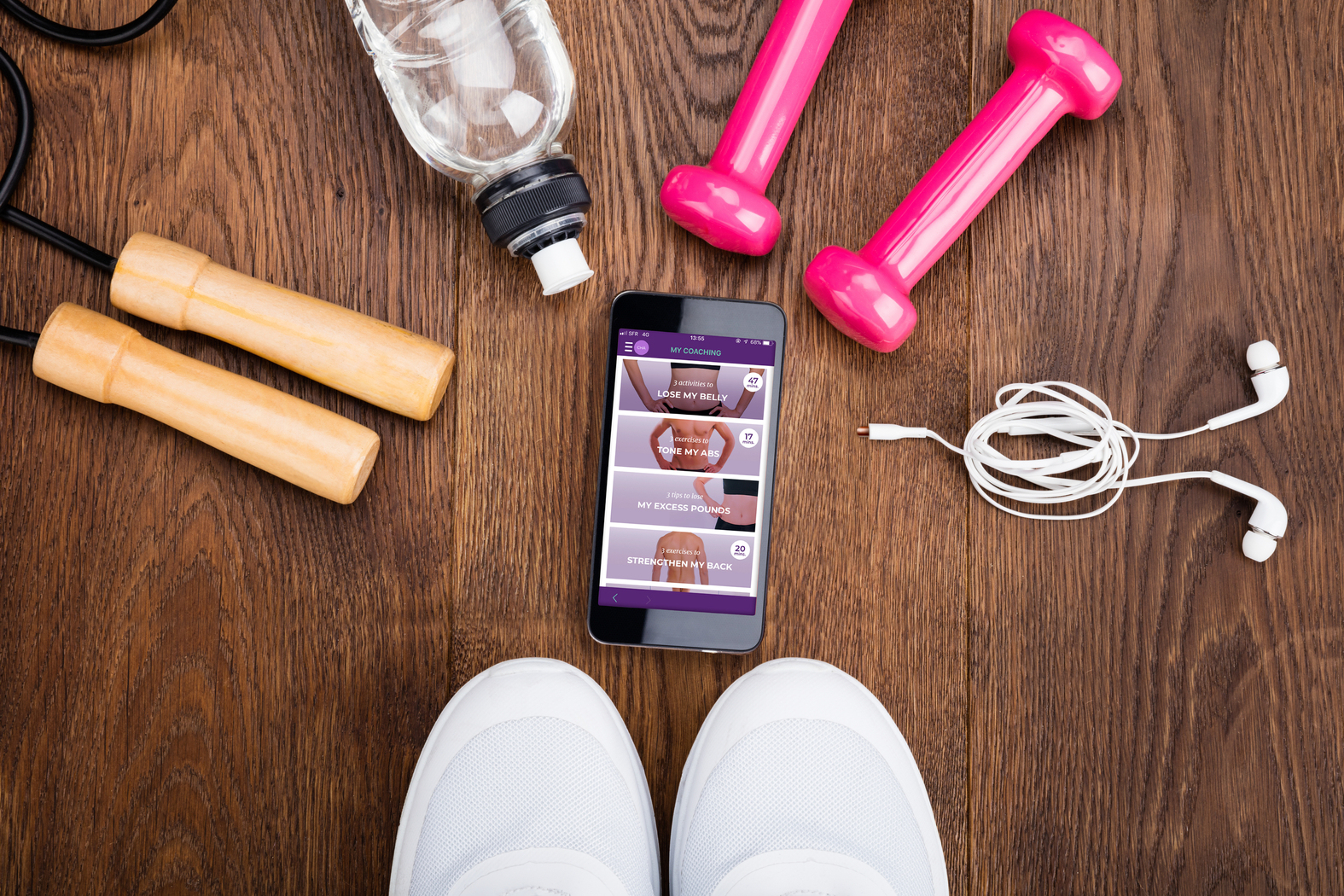 Sprzęt fitness na drewnianym tle: butelka wody, hantle, telefon, skakanka, trampki.