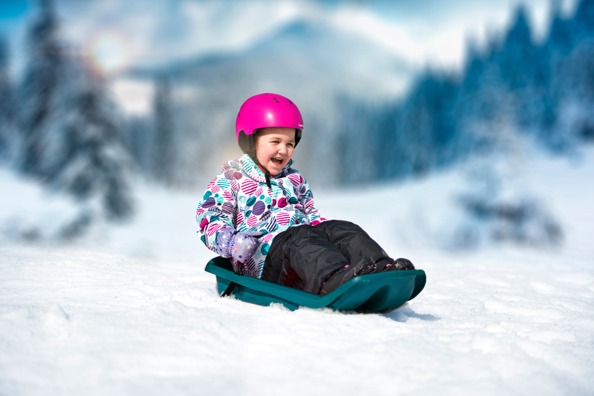 Dziewczyna w różowym kasku zjeżdża z ośnieżonego wzgórza, uśmiechając się. W tle ośnieżone góry.