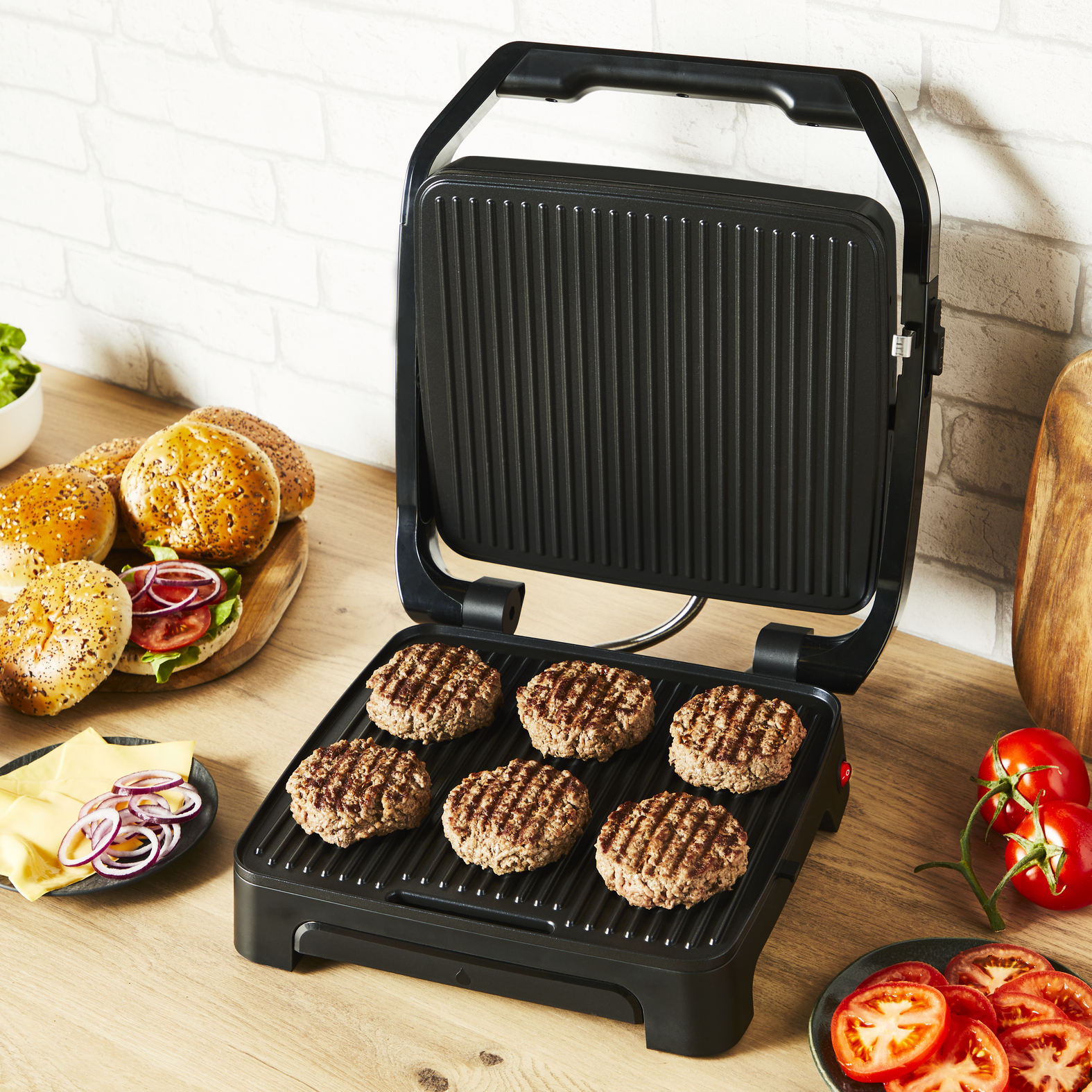 Burgery grillują się na czarnym grillu. Pokrojone pomidory, ser i cebula na blacie.