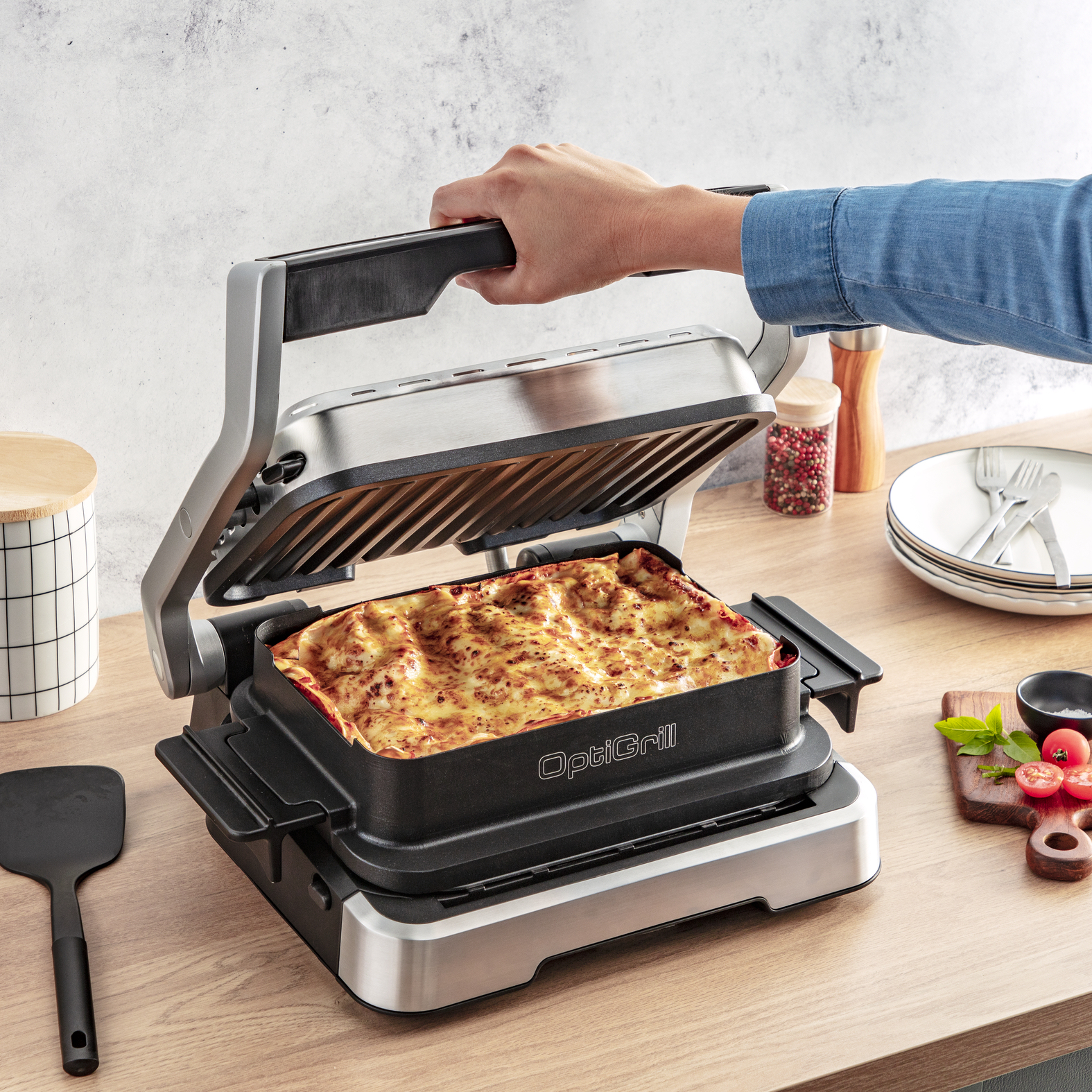 Eine Hand hebt den Deckel eines OptiGrills und zeigt eine Lasagne. Holztisch, Teller und Zutaten sind vorhanden.