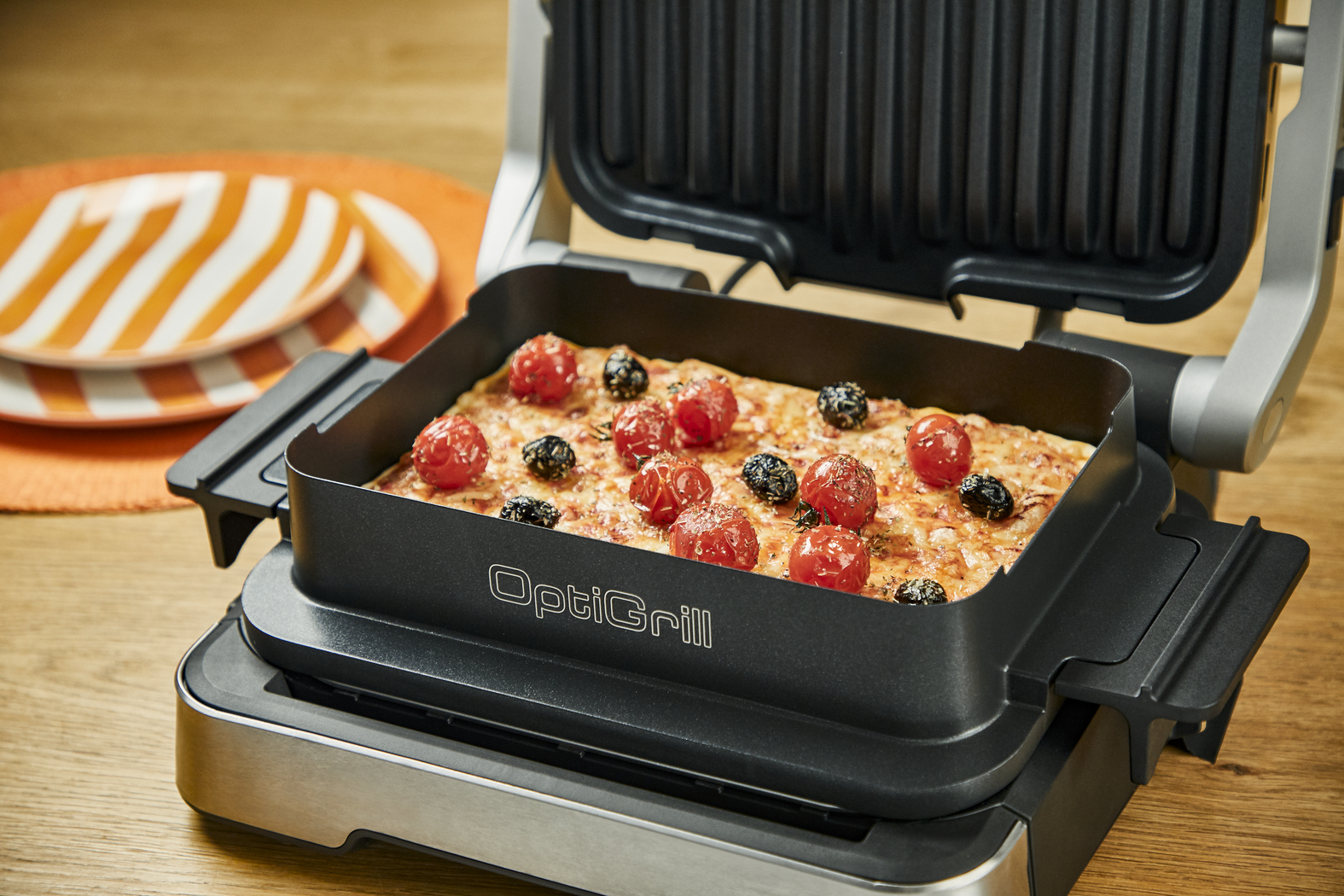 Ein OptiGrill mit Pizza, Tomaten und Oliven obenauf. Holztisch. Nahaufnahme.