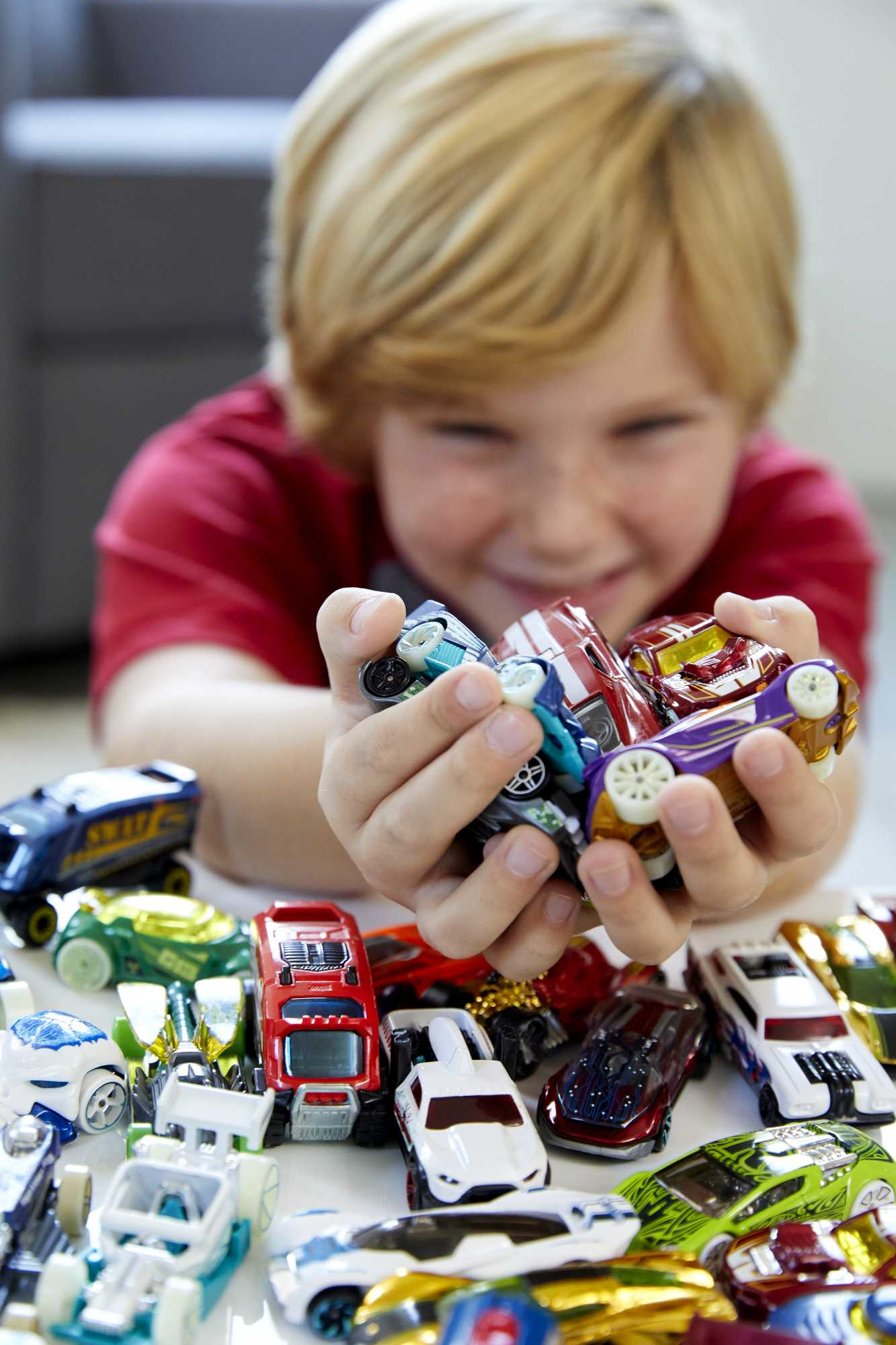 Ein junger Junge hält mehrere Spielzeugautos in seinen Händen, lächelnd, verschwommener Hintergrund.