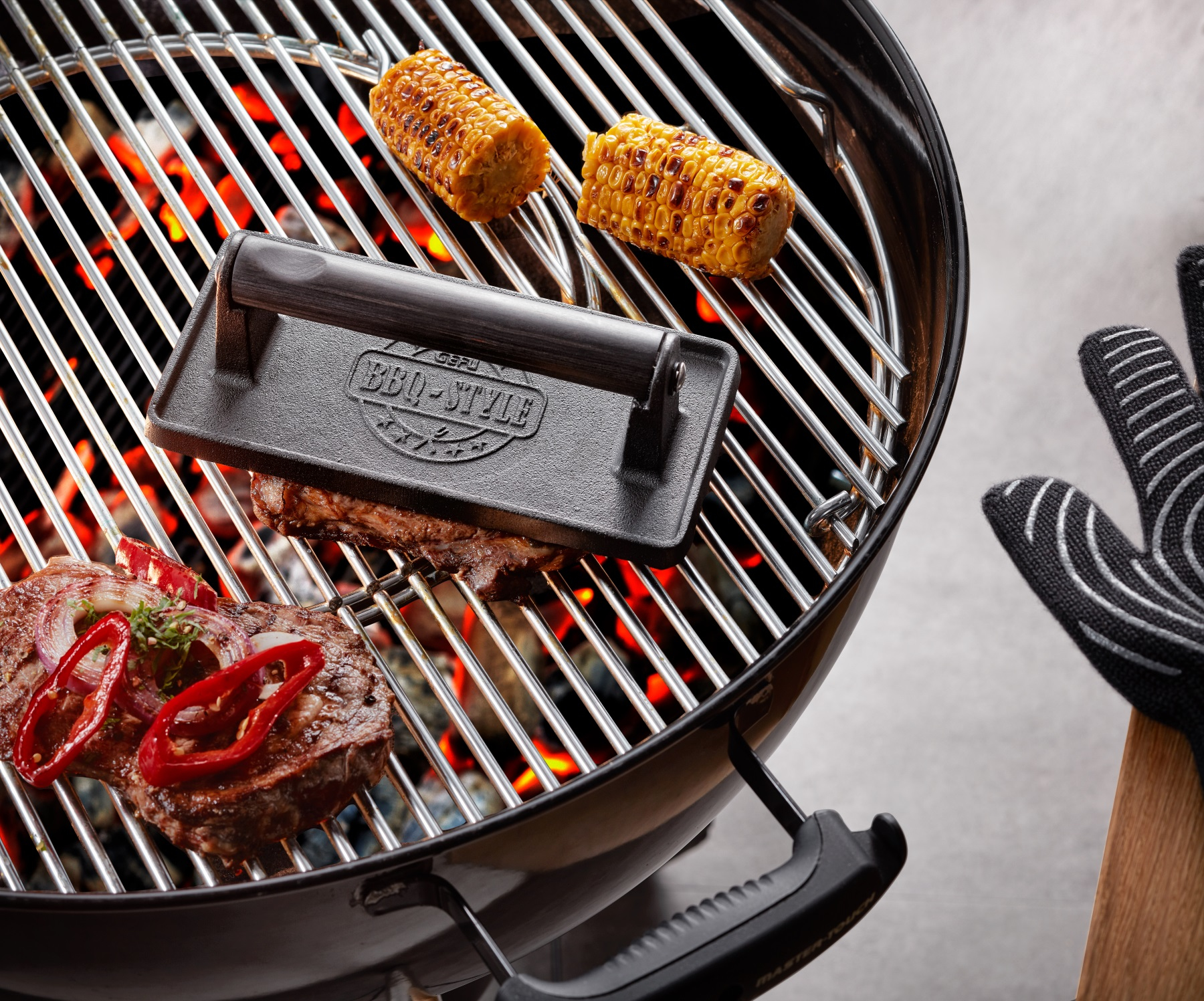 Grill z stekami, kukurydzą i prasą. Widoczne czerwone węgle. Czarne rękawice z boku.