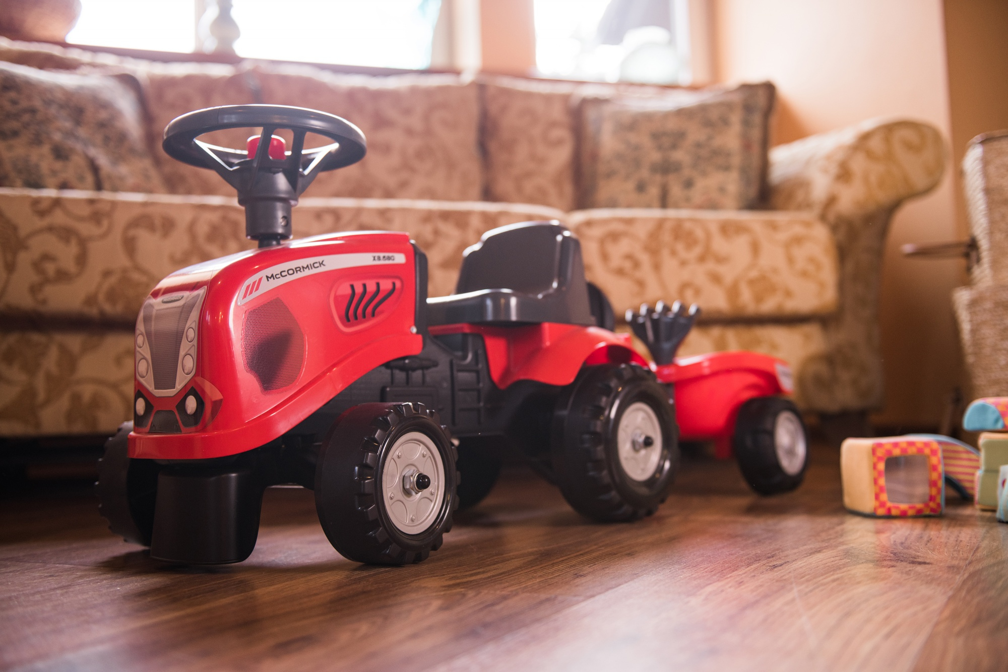 Tractor y remolque de juguete rojos, en suelo de madera, frente a un sofá beige.