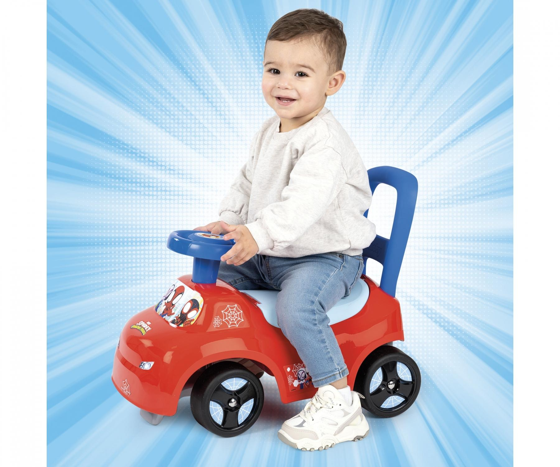 Ein Kleinkind sitzt lächelnd auf einem roten Spider-Man-Schiebewagen, mit blauem Hintergrund.