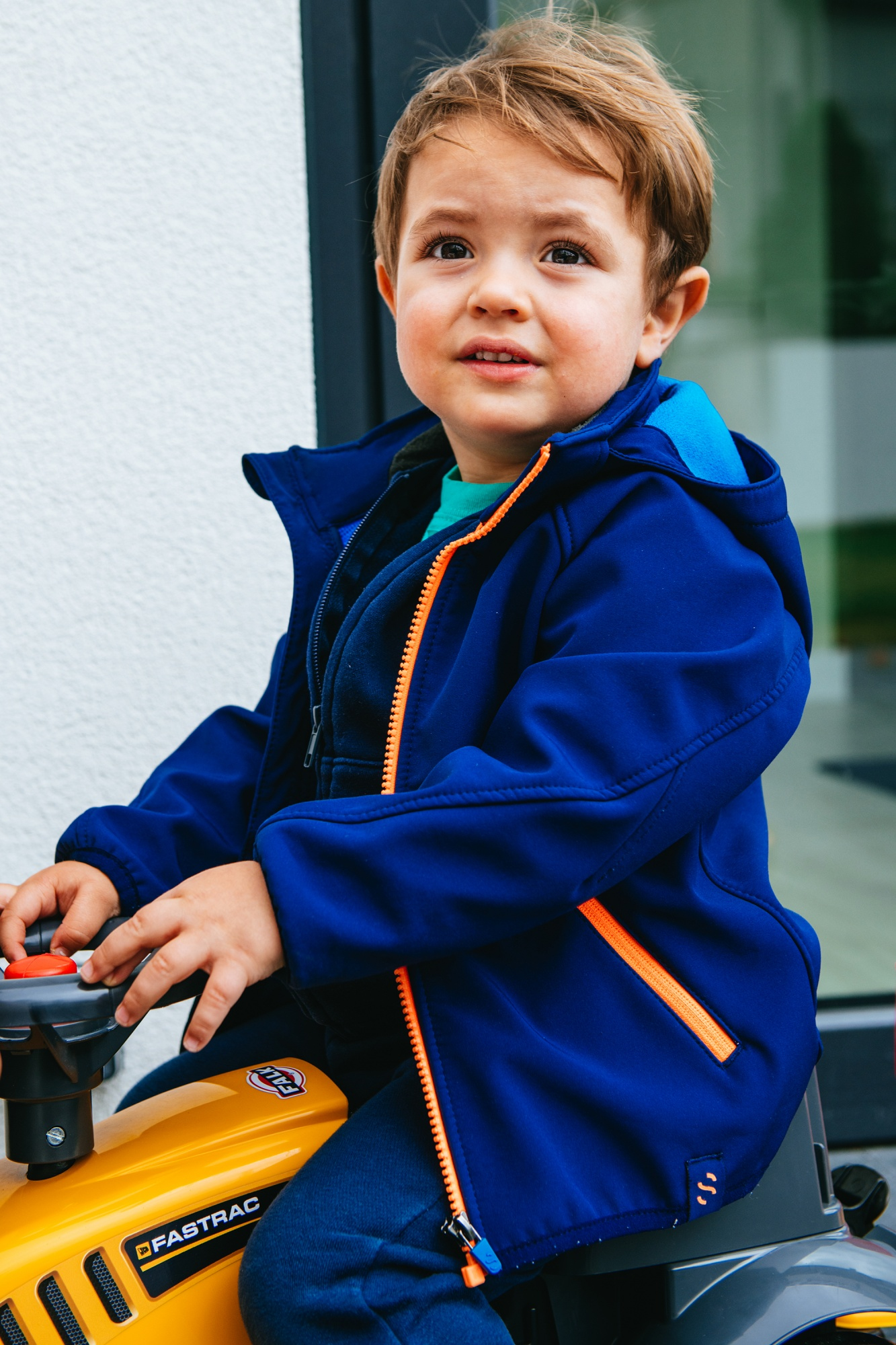 Un bambino con giacca e pantaloni blu seduto su un trattore giocattolo giallo.