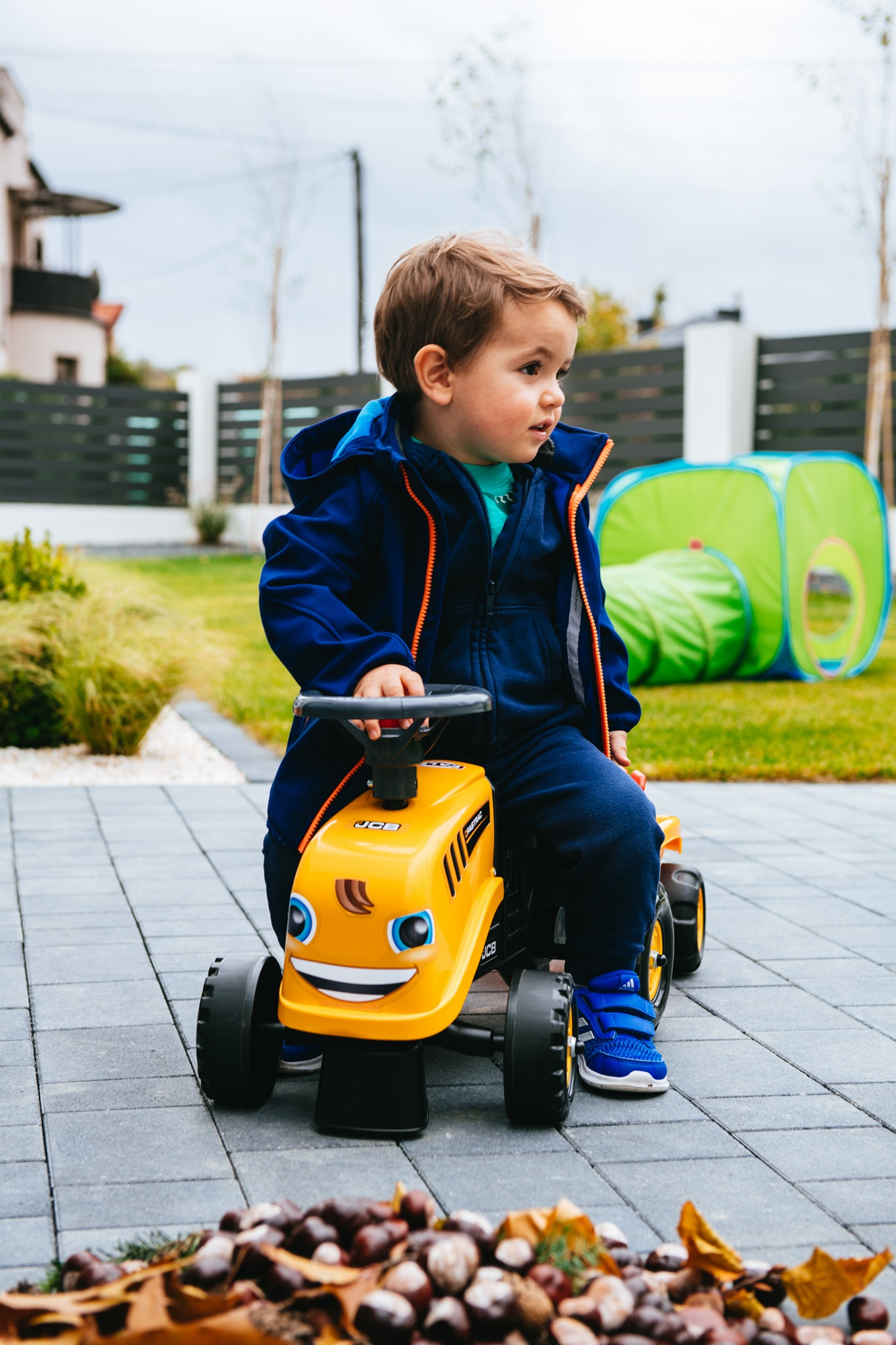 Un bambino su un trattore giocattolo giallo in giardino, indossa una giacca blu.