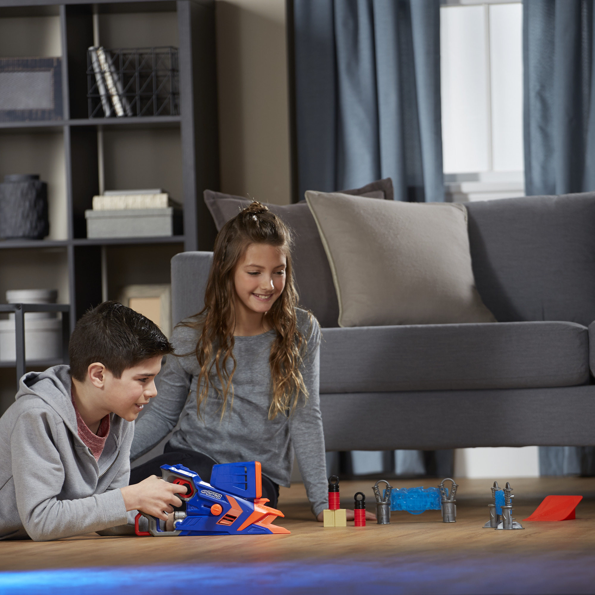 Dwoje dzieci bawi się pistoletem na zabawki i przeszkodami na podłodze. W tle sofa i półki.