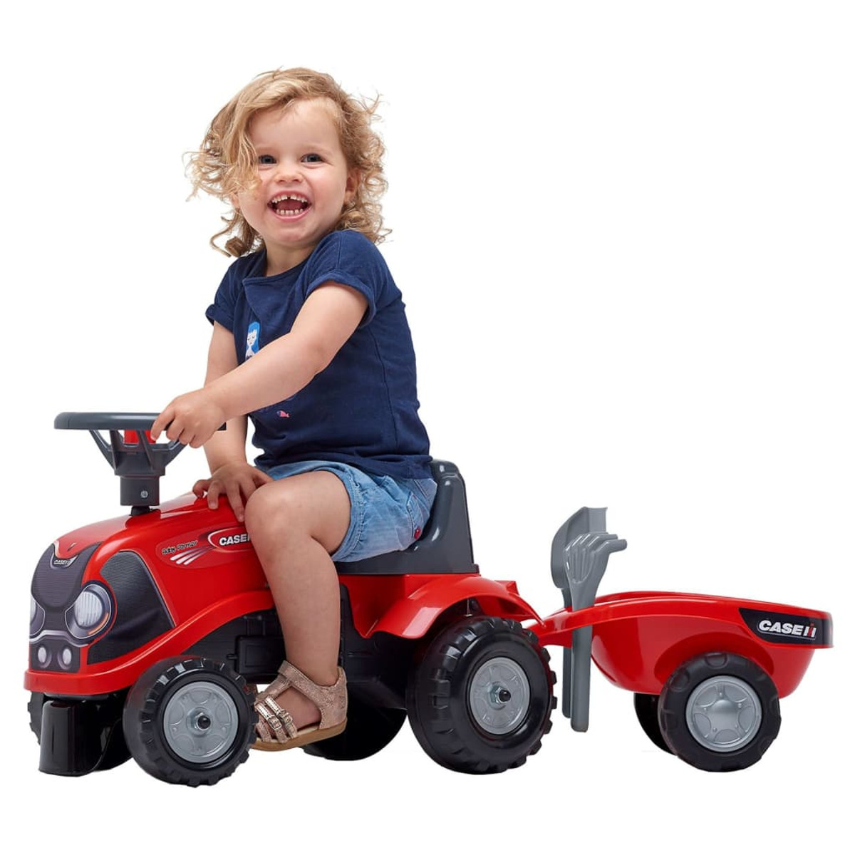 Una niña sentada en un tractor de juguete rojo con remolque, sonriendo y mirando a la cámara.