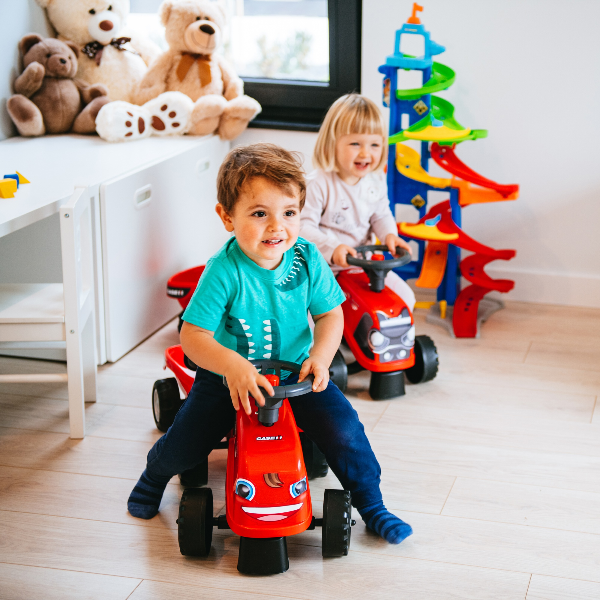 Dos niños, uno conduciendo un tractor rojo de juguete, el otro en un coche rojo de juguete, sonriendo.