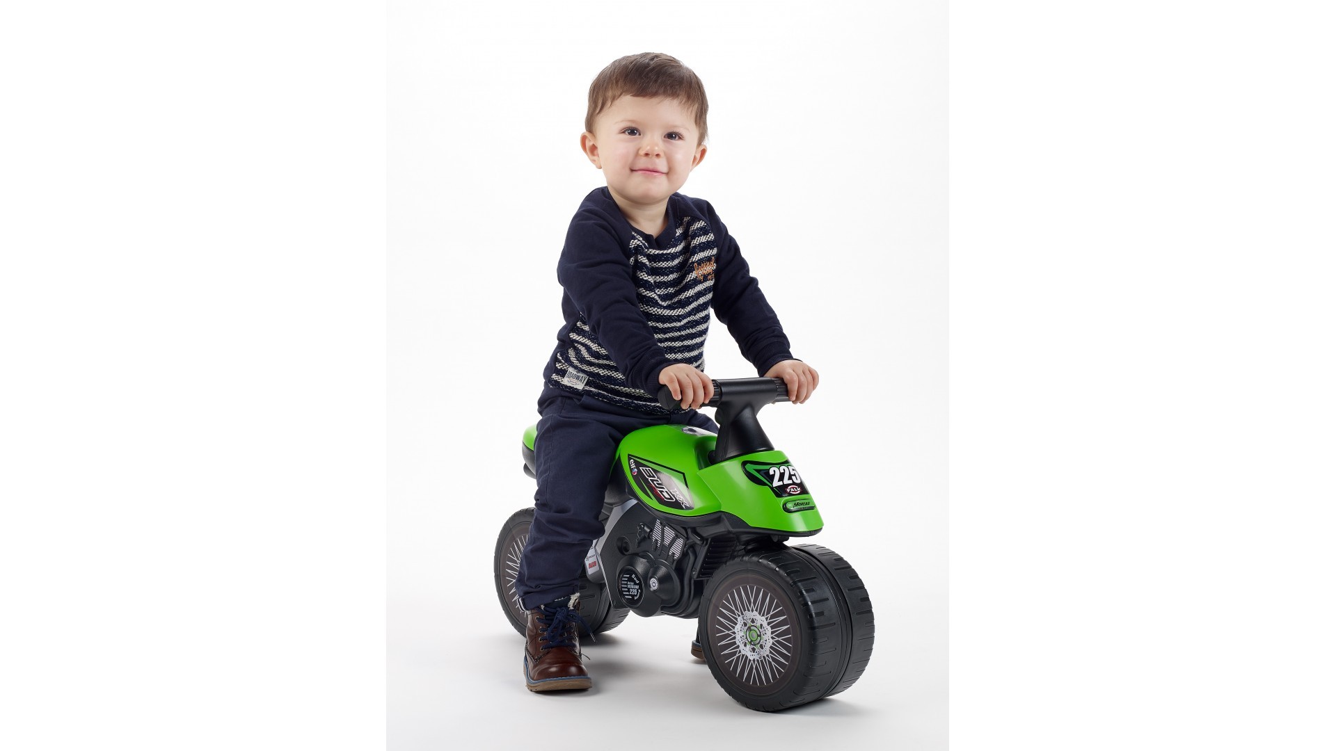 Bambino piccolo su una moto giocattolo verde. Il bambino ha una camicia a righe e sorride.
