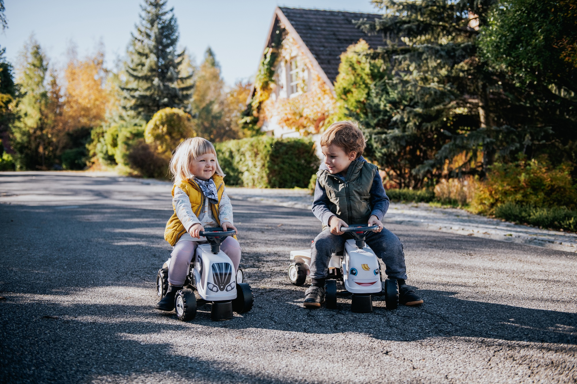 Zwei Kinder fahren mit Spielzeugtraktoren eine Straße entlang. Bäume und ein Haus im Hintergrund.