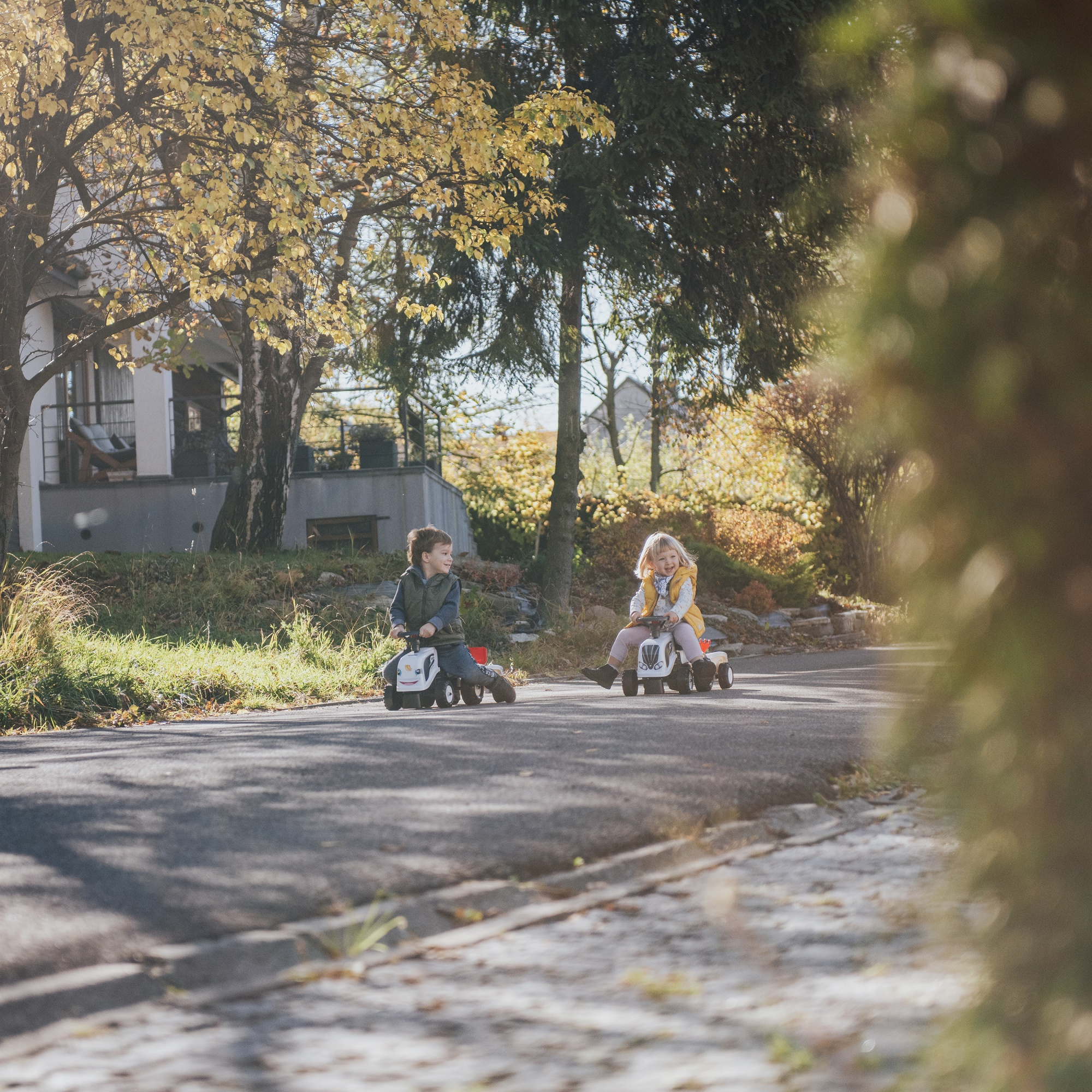 Zwei Kinder fahren auf Spielzeugautos auf einer Straße mit Bäumen und Häusern im Hintergrund.