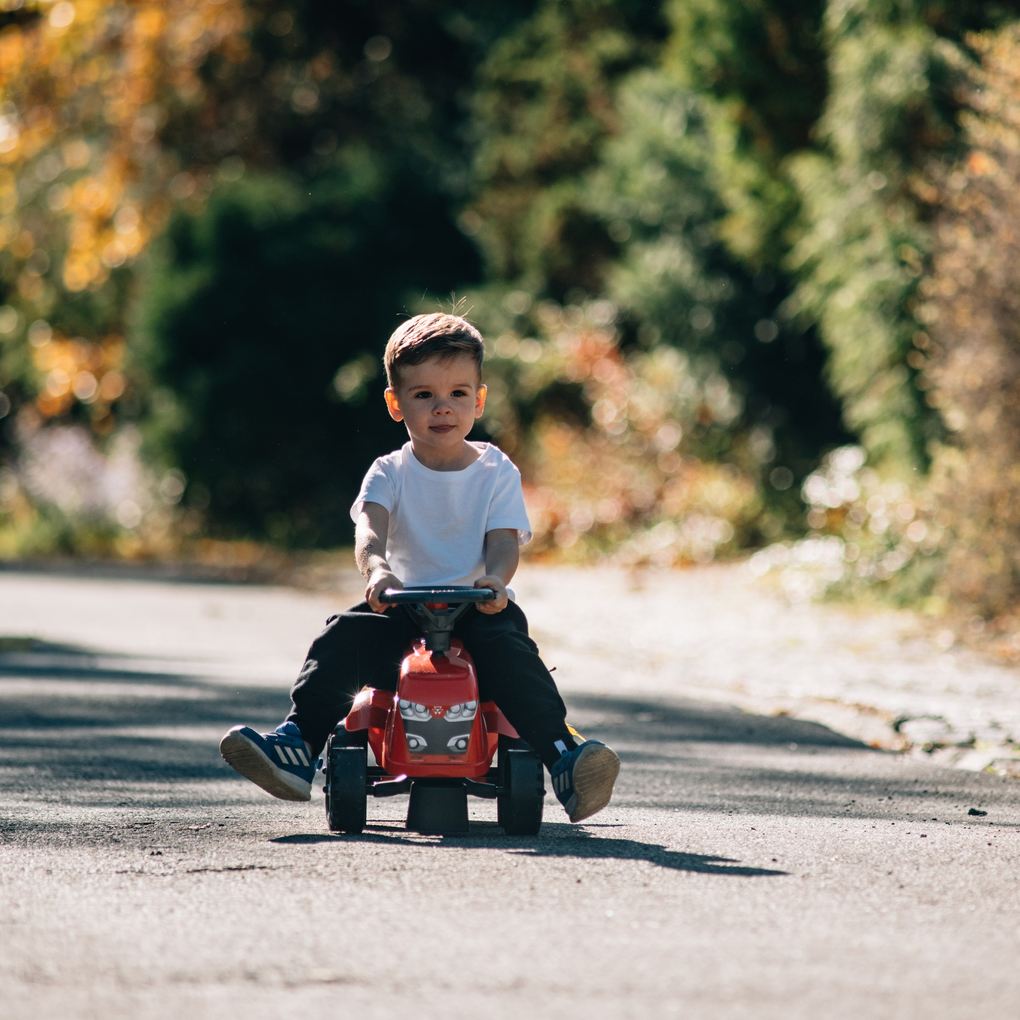Un niño pequeño en un tractor de juguete rojo en una carretera, sonriendo a la cámara.