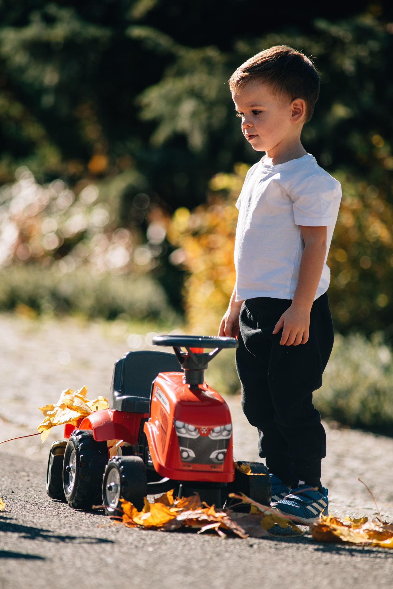 Un niño pequeño de pie junto a un tractor de juguete rojo con hojas caídas.