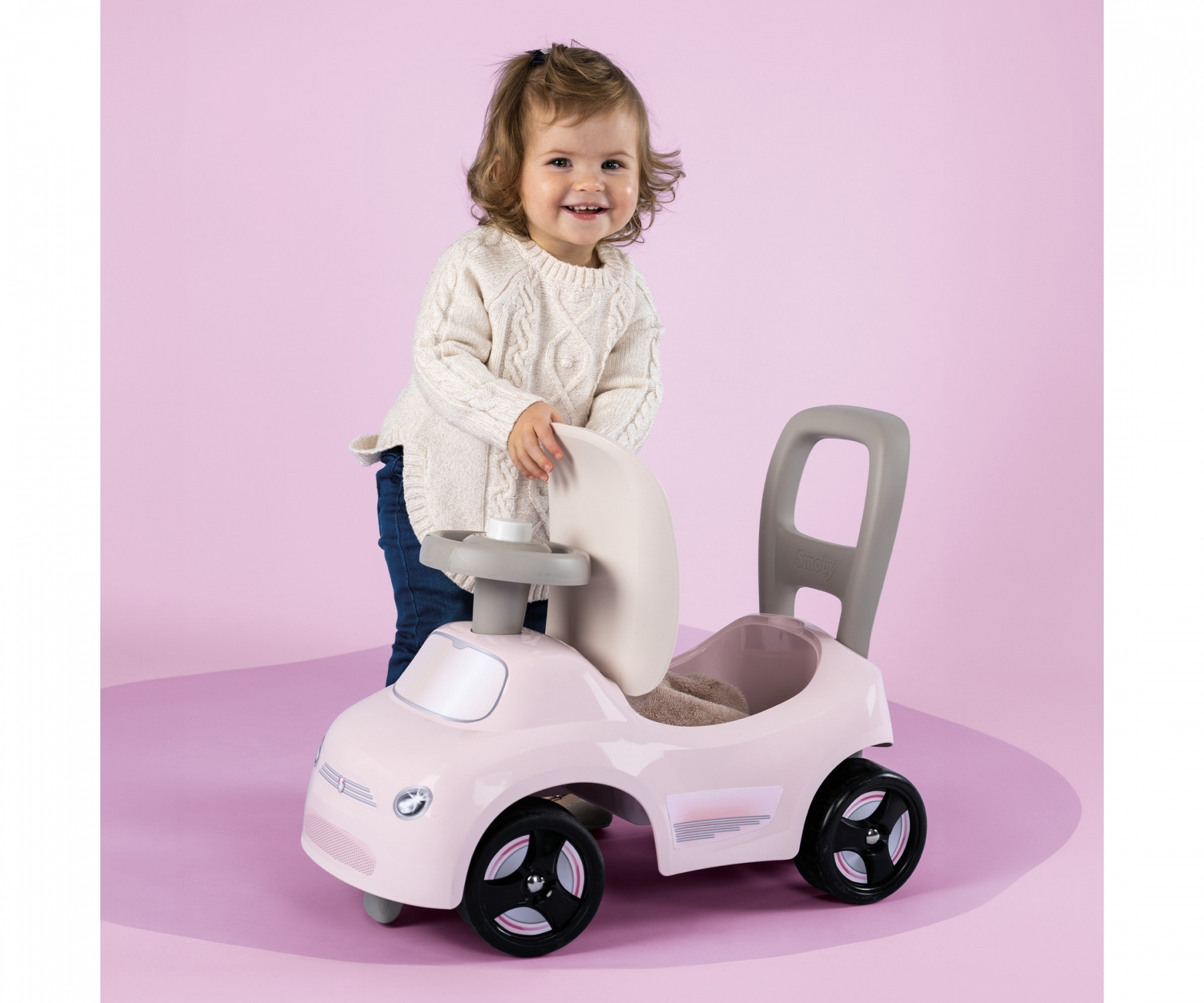 Małe dziecko stoi obok różowego samochodzika. Samochód ma uchwyt i kierownicę.