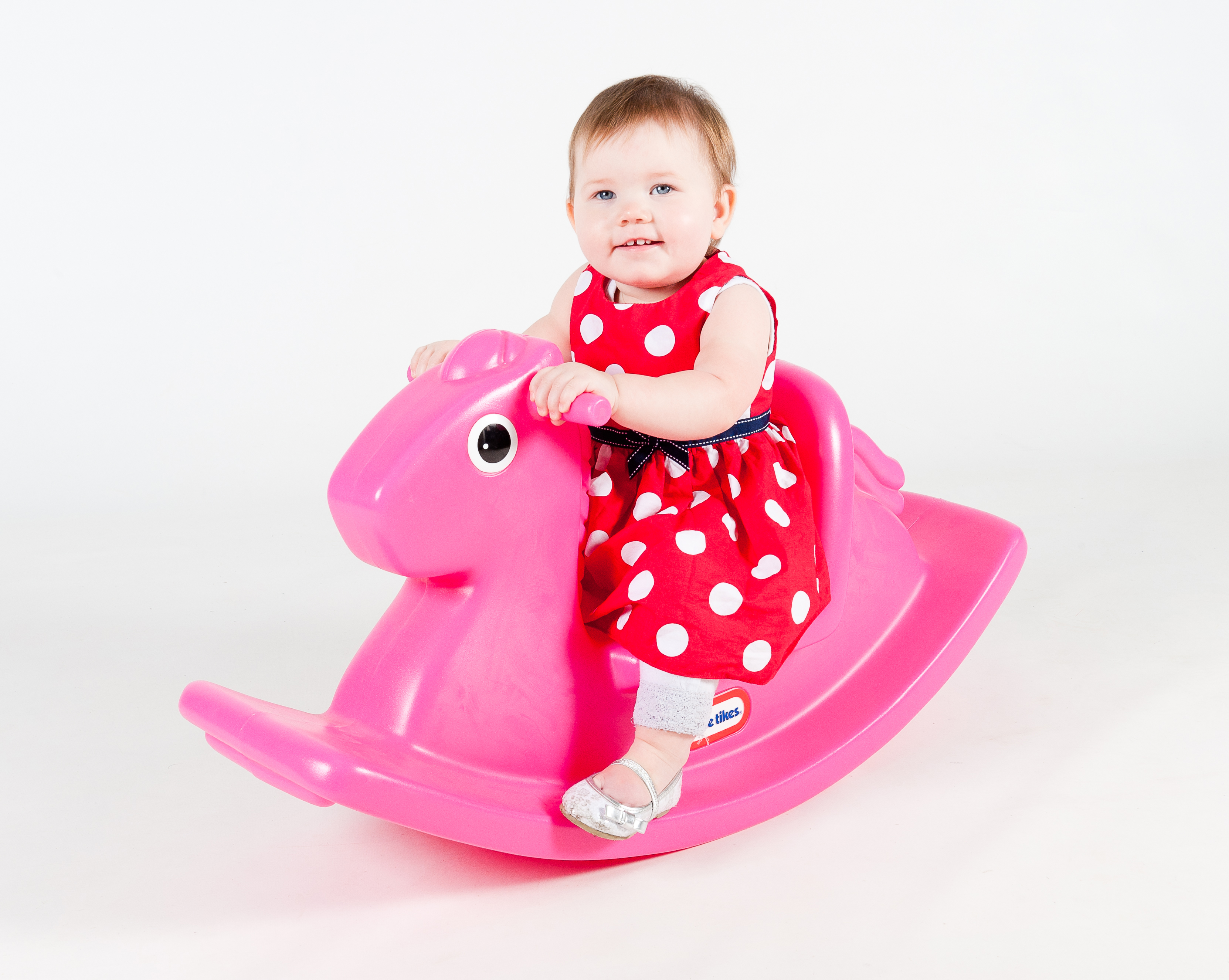 Bambina su un cavallo a dondolo rosa, indossa un vestito rosso a pois bianchi. Sfondo bianco.