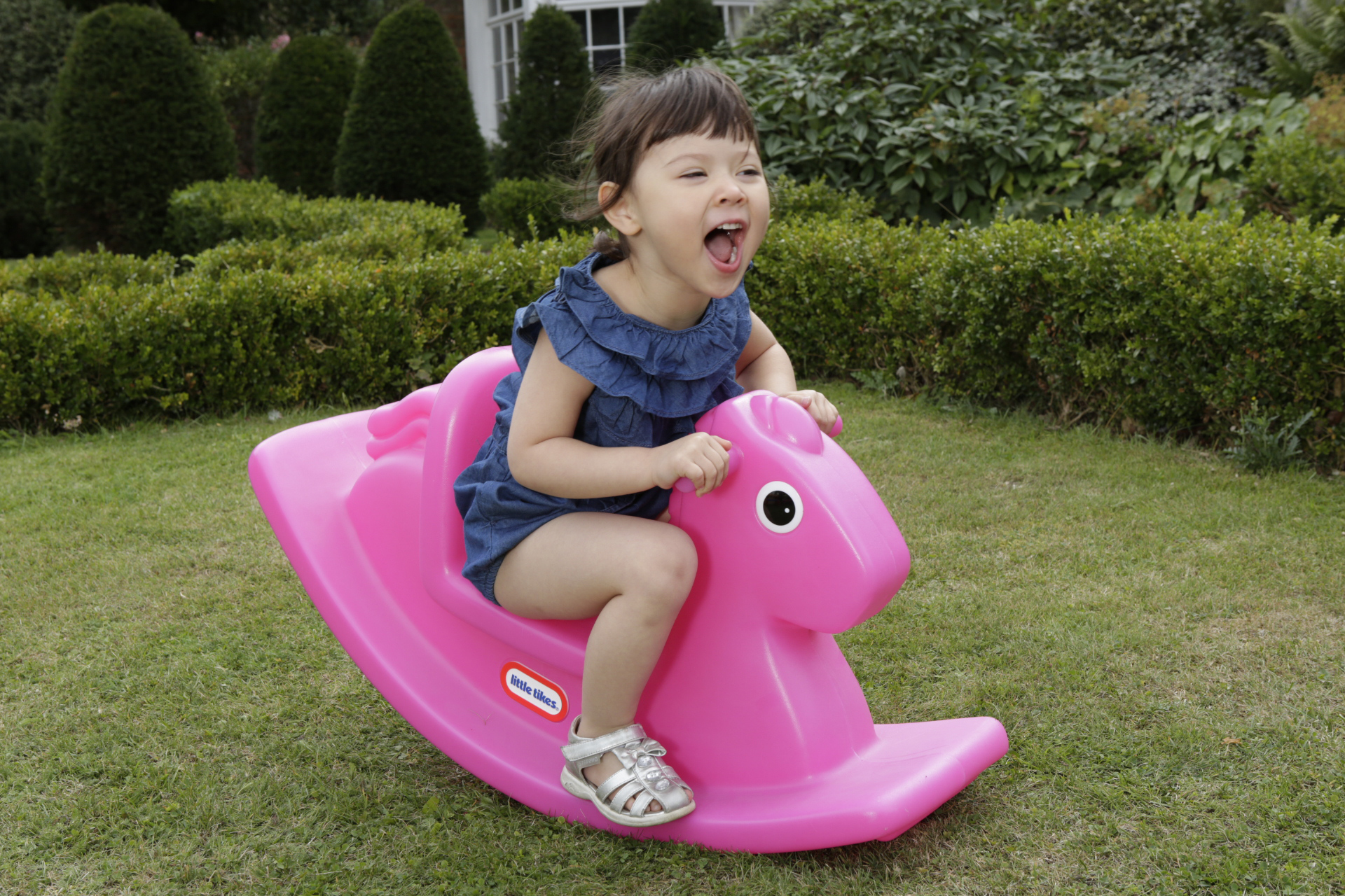 Una bambina sorride mentre cavalca un cavallo a dondolo rosa su un prato verde.