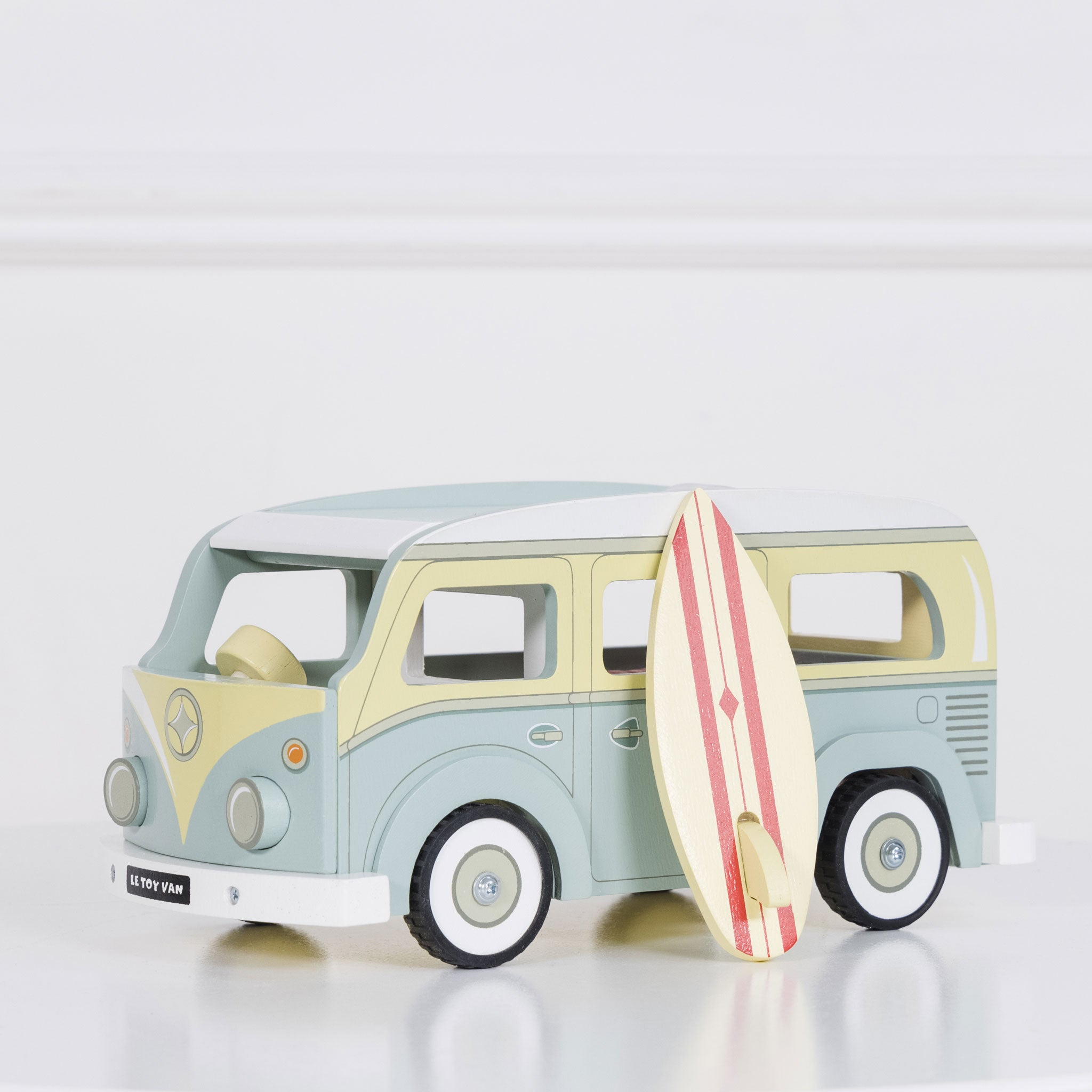 Ein Holzspielzeug-Van, hellblau und gelb, mit einem Surfbrett, das daran lehnt. Der Van steht auf einer weißen Oberfläche.