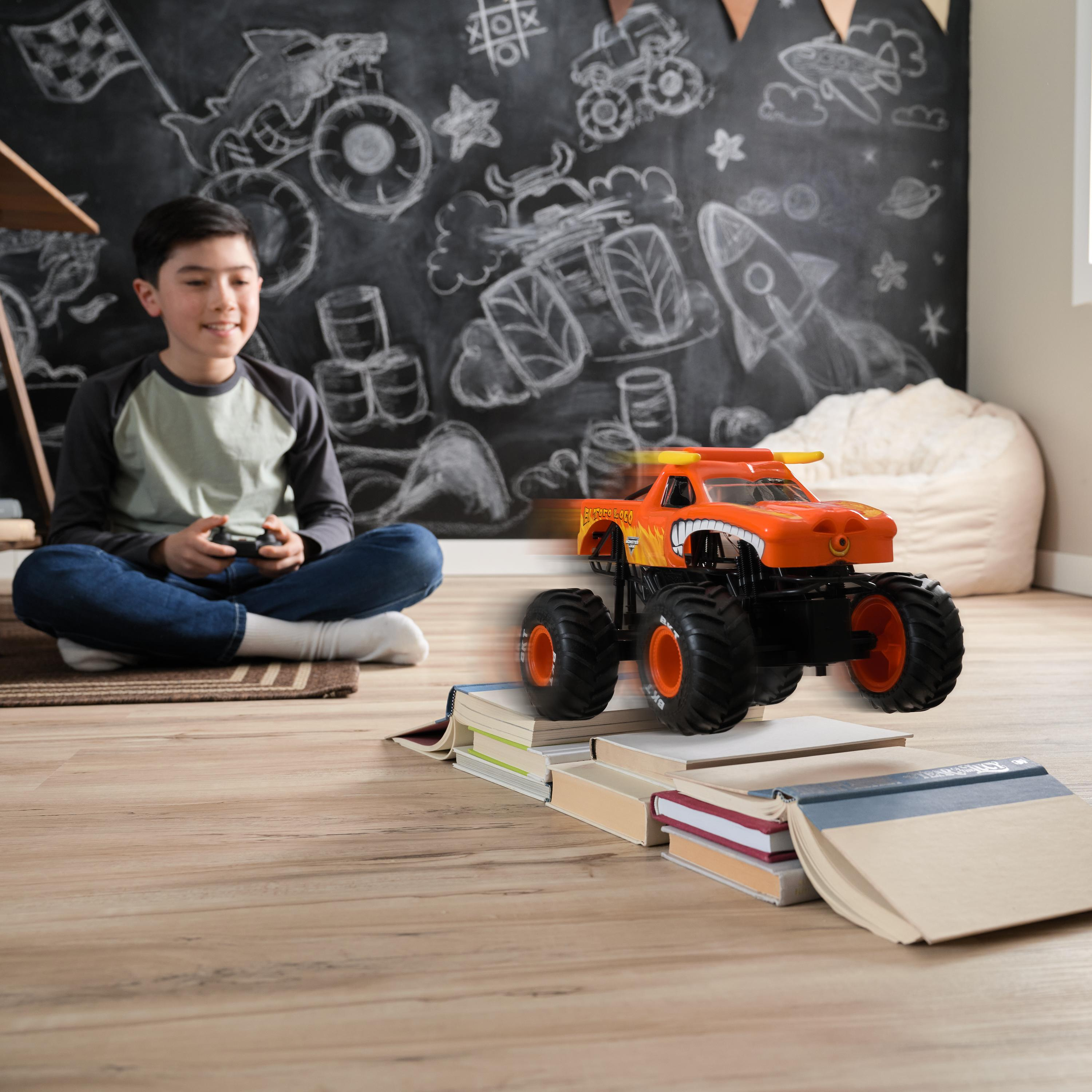 Chłopiec bawiący się zabawką monster truck, jadący po książkach na drewnianej podłodze. Ciężarówka jest pomarańczowa.