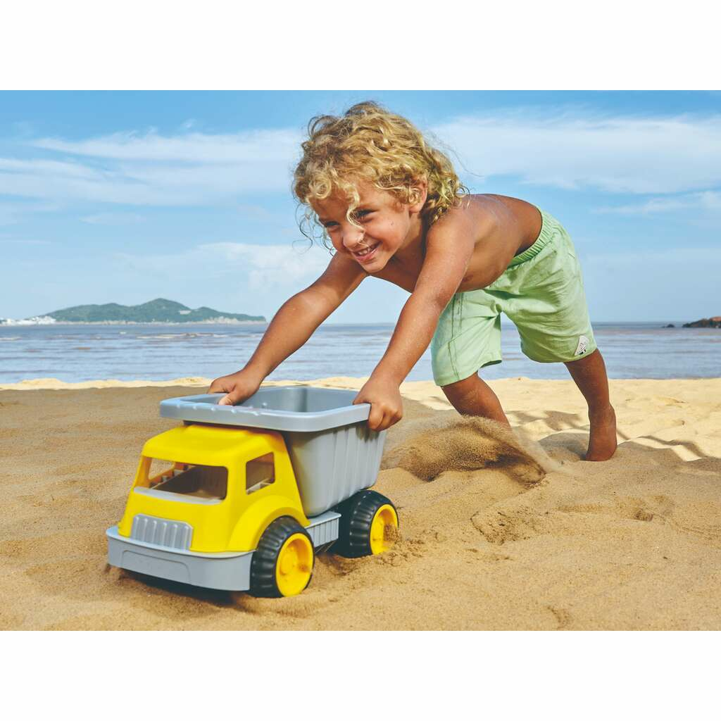 Un ragazzino spinge un camioncino giallo e grigio su una spiaggia sabbiosa. Cielo blu e acqua sullo sfondo.