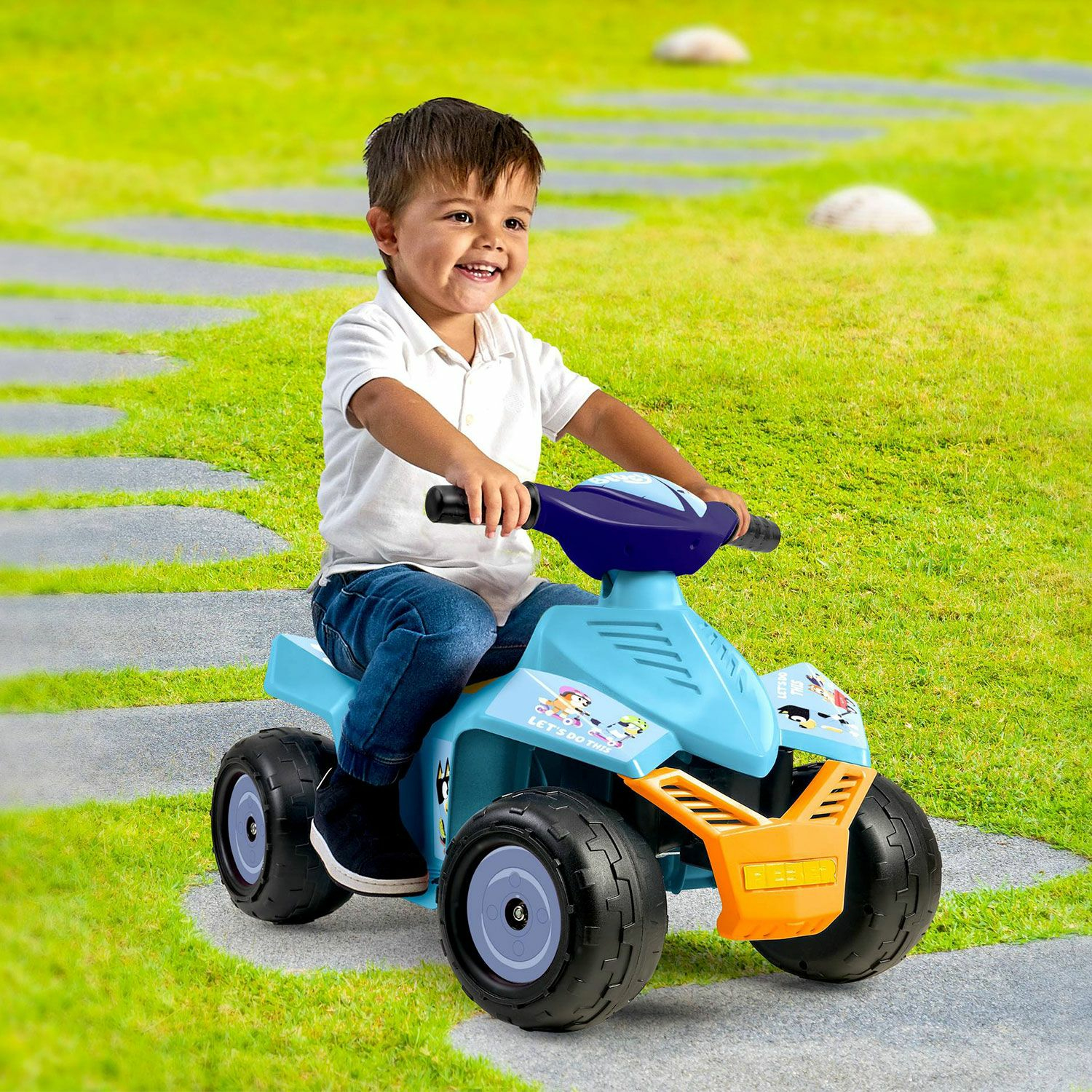 Ein Junge fährt auf einem blauen und orangefarbenen Spielzeug-ATV auf einem Steinweg mit Gras.