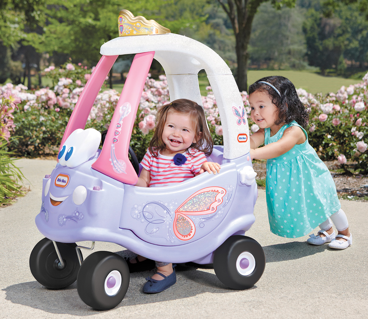 Due bambine giocano con un'auto giocattolo. Una è dentro, l'altra la spinge. Auto rosa, viola e bianca.