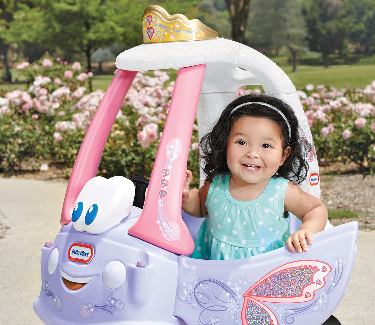 Una bambina è seduta in un'auto giocattolo viola con una corona e un design a farfalla. Lo sfondo è un giardino.