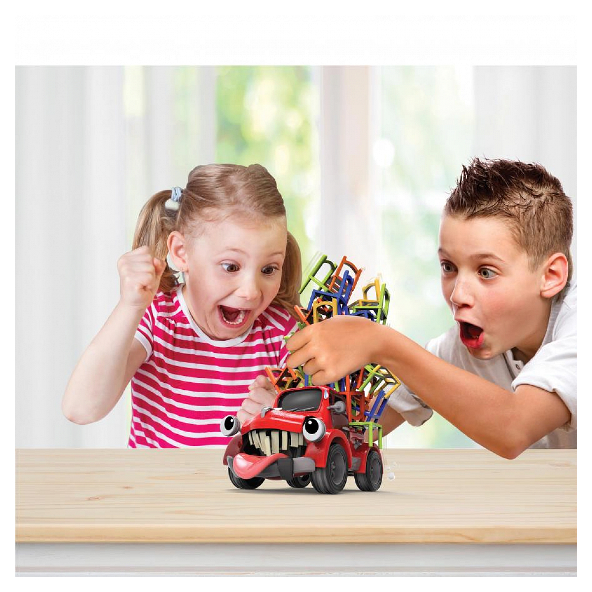 Dwoje dzieci z ekscytacją patrzy na czerwoną ciężarówkę z zabawkami równoważącą kolorowe krzesła.