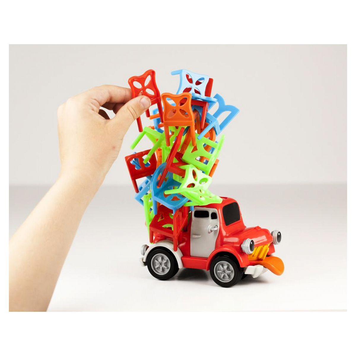 Czerwona zabawka ciężarówka z kolorowymi krzesłami na górze i ręka sięgająca po krzesła.