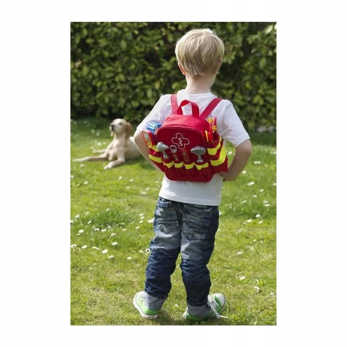 Chłopiec z czerwonym plecakiem medycznym, z psem leżącym na trawie w tle.