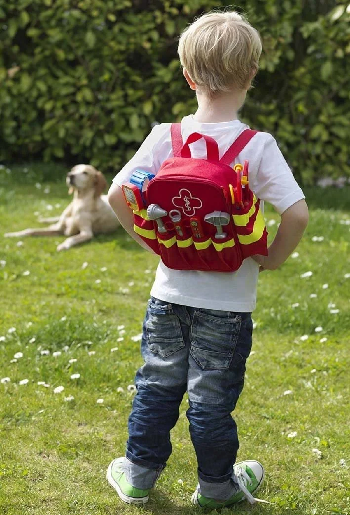 Chłopiec w białej koszuli i dżinsach, noszący czerwony plecak z zabawkami. Pies leży w tle.